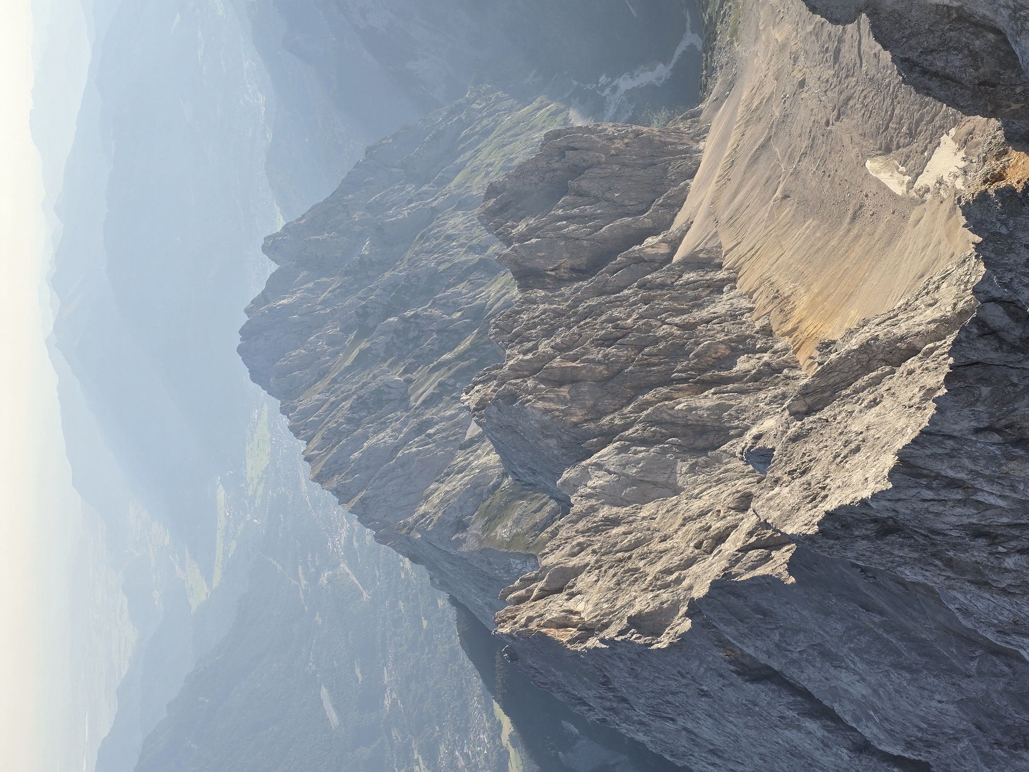 This is a high-angle, full shot of a rugged mountain range in Grainau, Germany. The foreground is dominated by a steep, rocky ridge with sharp, jagged peaks. The rock is a light gray and shows clear signs of erosion and fracturing. The ridge slopes down to the right, revealing a barren, sandy slope littered with debris and scree.
Beyond the immediate foreground, the mountain range stretches into the distance, with successive ridges fading into a hazy, blue-gray atmosphere. The distant mountains appear softer and less defined, creating a sense of depth and scale. In the far background, a valley with a winding river and scattered settlements is visible, appearing as a muted green and brown patchwork. The sky is bright and clear, with a subtle haze that softens the distant landscape. There are no people or animals visible in the image. The overall impression is one of a vast, natural, and somewhat imposing mountain environment.
