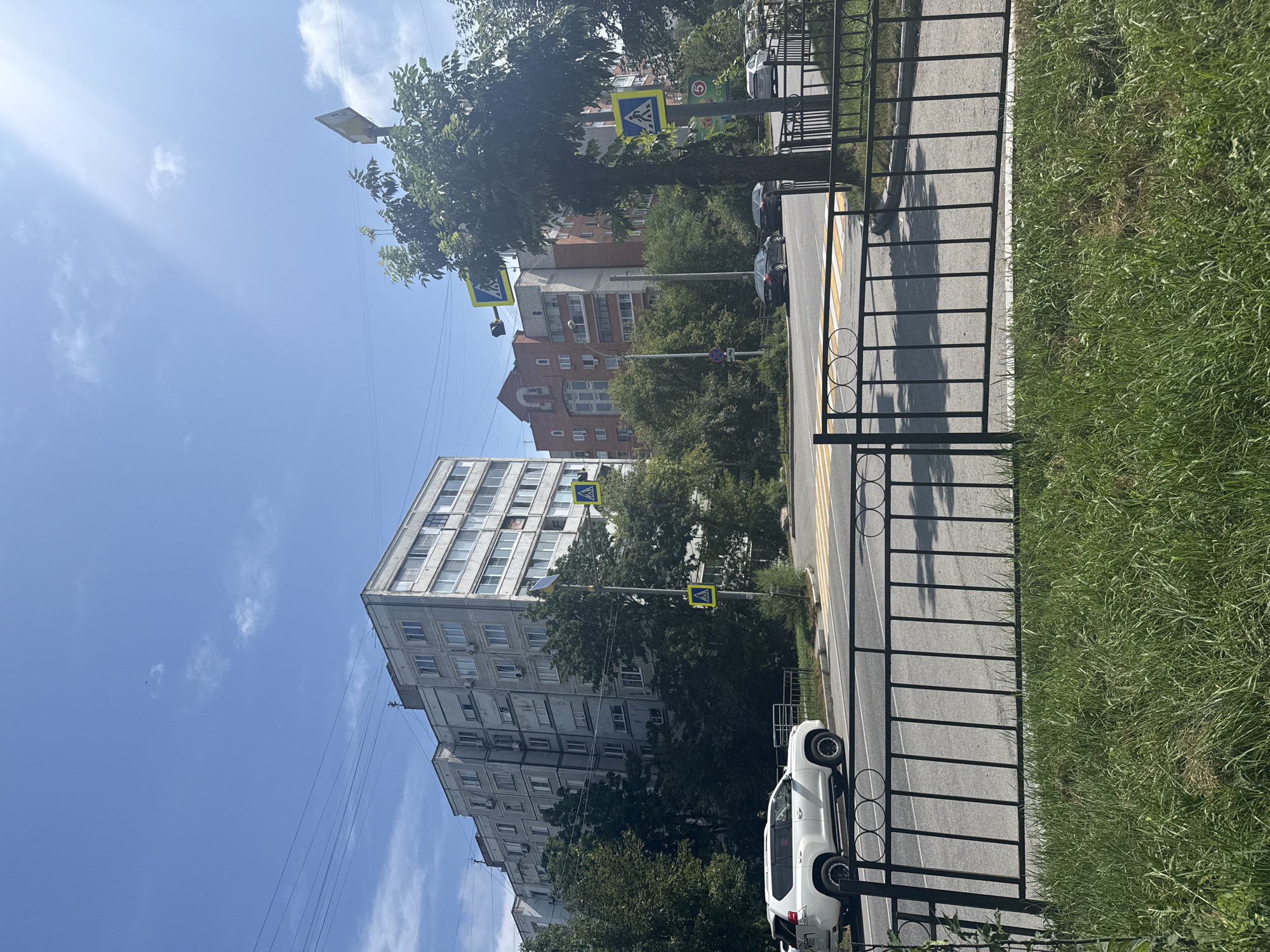 A street scene in Khabarovsk, Russia, on a sunny day with a bright blue sky and scattered clouds. In the foreground, a black metal fence separates a grassy verge from a paved road with a yellow and white striped pedestrian crossing. A white SUV is parked on the right side of the road, facing left. Beyond the pedestrian crossing, several multi-story apartment buildings are visible, characterized by their utilitarian architecture. One building on the left is grey and appears to be of Soviet-era construction, while a taller, brick-colored building with a distinctive gabled roof is situated further back. Trees line the street, with dense green foliage in front of the buildings. Pedestrian crossing signs with yellow triangular backgrounds and black silhouettes of walking figures are posted on poles at various points along the road. There are no people actively crossing the street or interacting with each other. The overall impression is a typical urban streetscape in a Russian city.