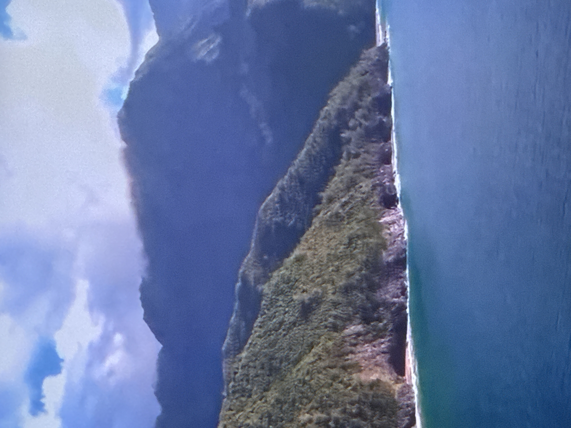 A vast expanse of calm, blue ocean dominates the foreground, meeting a rugged coastline. Lush green, tree-covered hills rise steeply from the water, dotted with rocky outcrops and occasional dark cave openings. In the distance, imposing, dark mountains shrouded in mist and clouds form a dramatic backdrop. The sky above is a mixture of blue patches and soft, gray clouds, some tinged with pink light. The scene depicts a remote, natural landscape.