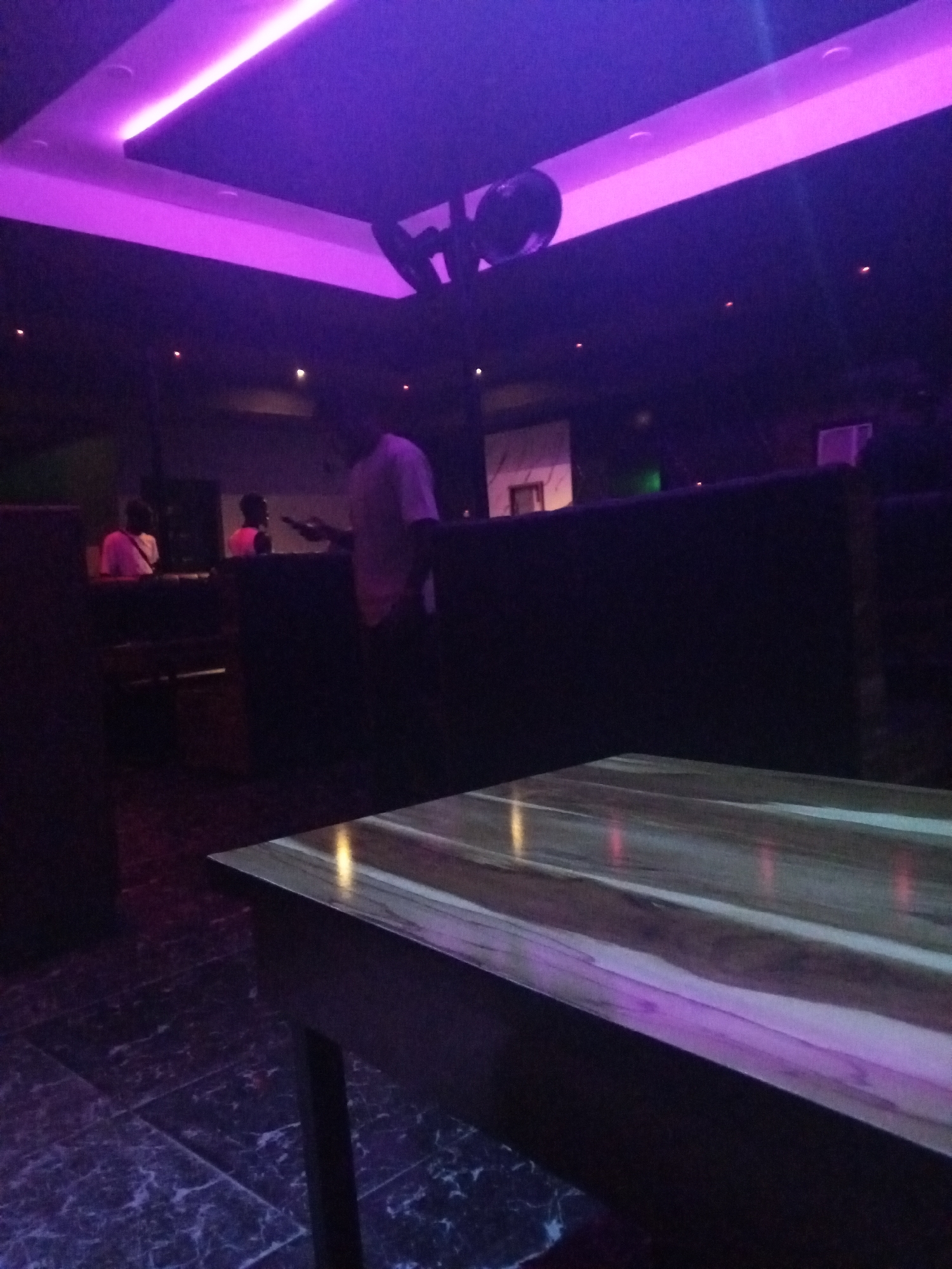 The image shows the interior of a bar or nightclub in Lokoja, Nigeria. The lighting is predominantly purple. A table is in the foreground. In the background, there are people standing near a bar or counter. A person is holding a phone. The floors appear to be tiled with a marble-like pattern.