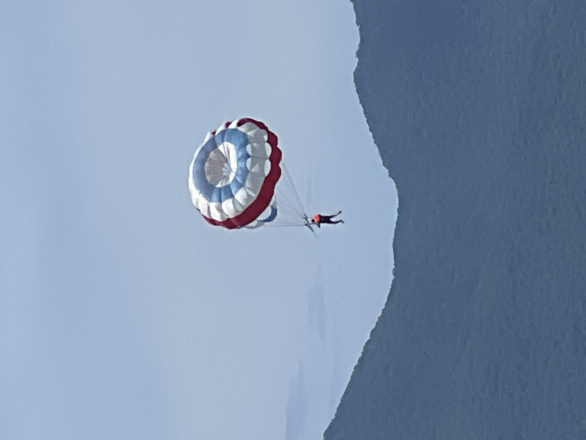 A person is parasailing high in the sky above the misty, dark blue mountains near Da Nang, Vietnam. The parasail is a large, circular canopy with alternating red, white, and blue sections. The person, wearing a dark outfit and an orange life vest, is suspended below the parasail by ropes, with their legs dangling in the air. The background is a clear, pale blue sky, with the jagged outline of mountains visible below, suggesting a coastal or island landscape. The image captures a moment of aerial leisure activity against a natural backdrop.