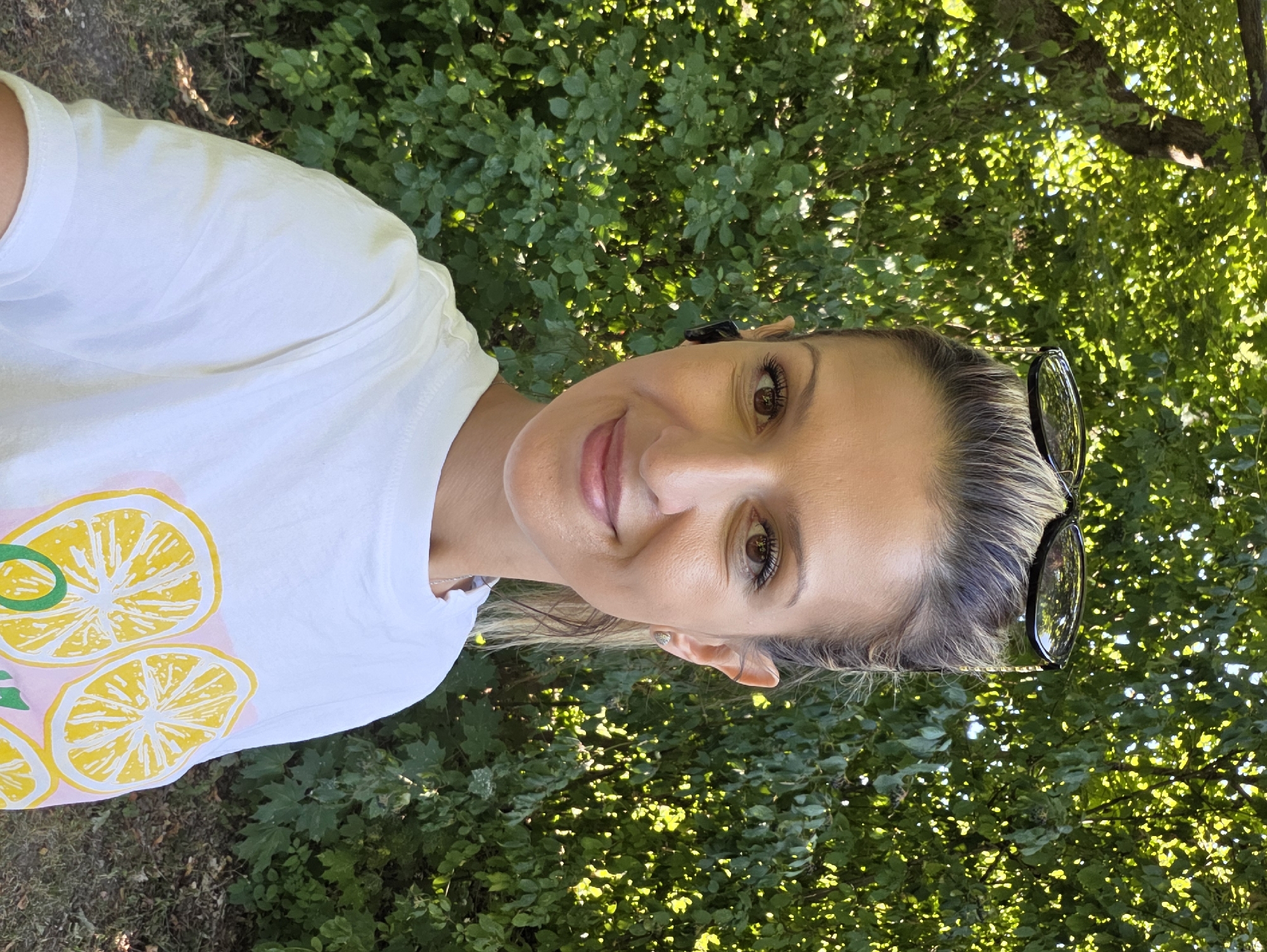 A young woman with blonde hair pulled back, sunglasses perched on top of her head, and wearing a white t-shirt with a lemon print, poses for a selfie outdoors. She is smiling and looking at the camera. The background is a lush green foliage of trees and bushes. The lighting suggests it is daytime, with sunlight filtering through the leaves. The location is not specified in the image.