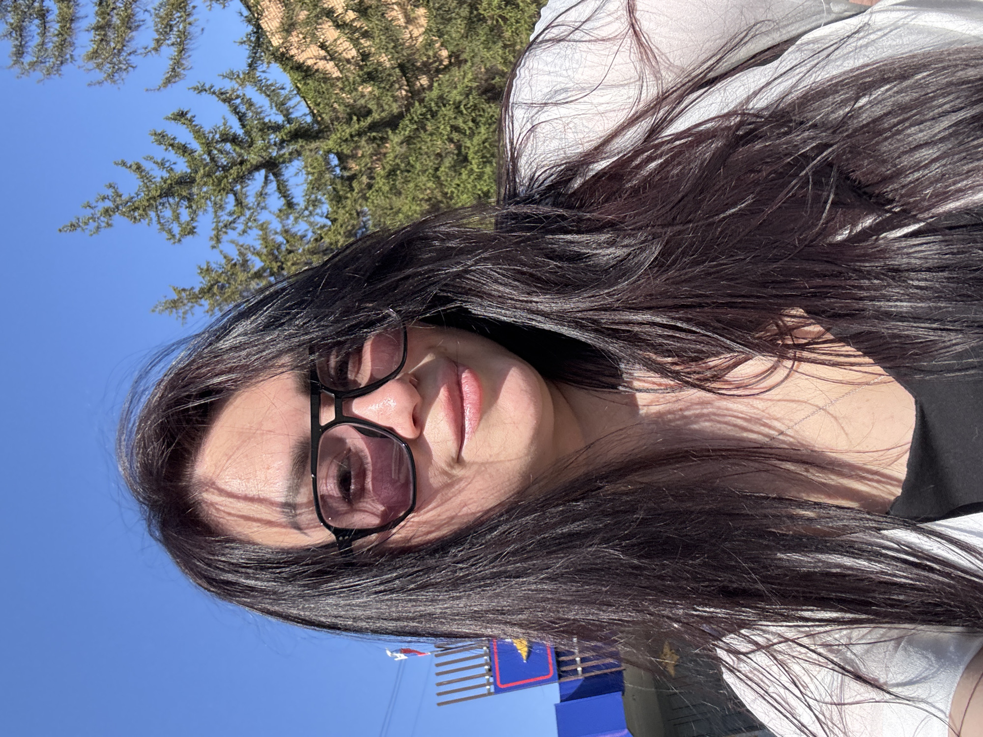 A selfie taken from a low angle shows a woman with long, dark hair and sunglasses. She wears a white top and is likely outdoors on a sunny day, judging by the bright blue sky and the sunlight on her face. In the background, there is a tree with dense green foliage and a blue and red structure, possibly part of an amusement park or other recreational facility. The location is inferred to be in the city of Unknown, Unknown.