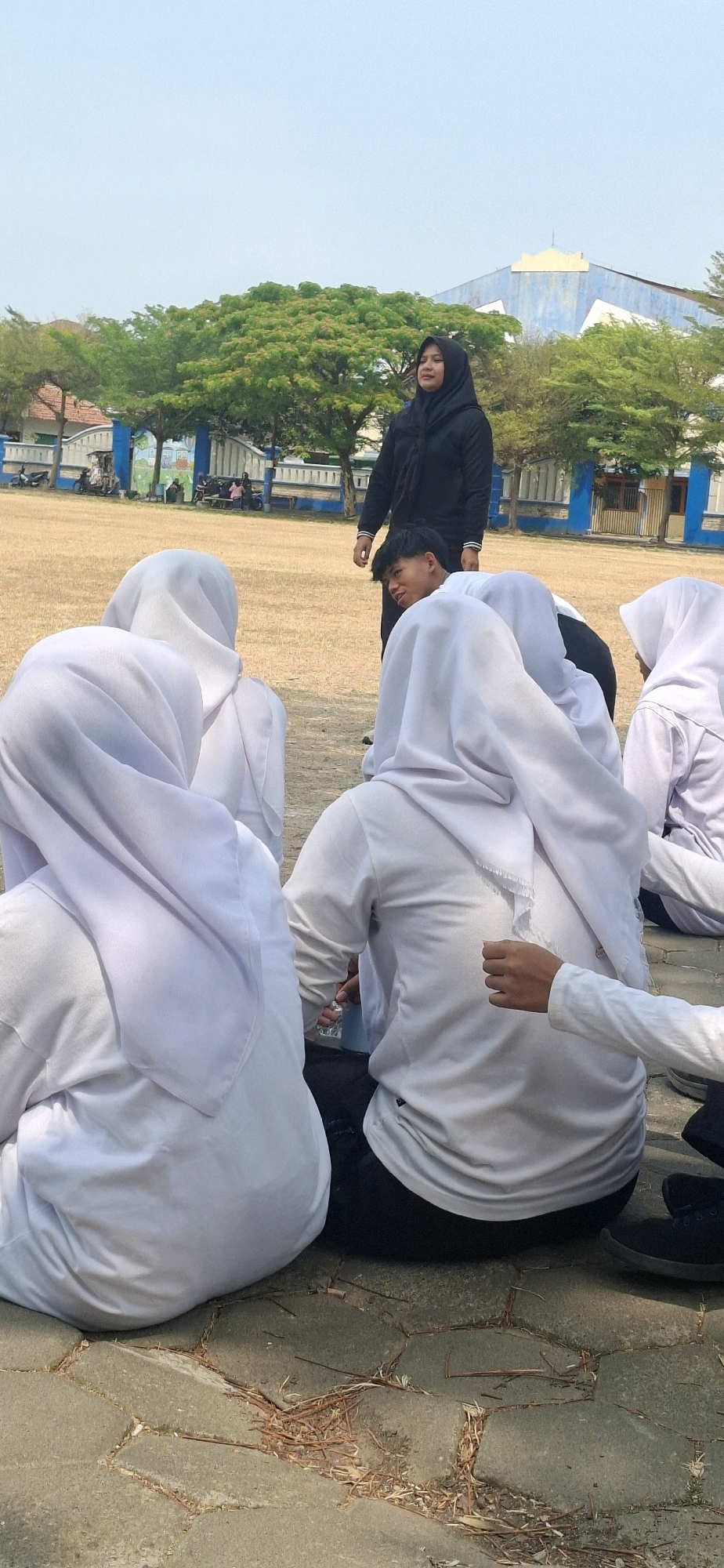 In Baturan, Indonesia, a group of individuals, predominantly young women wearing white hijabs and long-sleeved shirts, are seated on the ground in a paved outdoor area. Some are facing away from the camera, showing the backs of their heads and torsos. A woman in a black hijab and long-sleeved top stands behind them, looking towards the seated individuals. A young man in a white shirt is also present, turned slightly towards the camera with an expression of curiosity. The environment appears to be a field or park, with trees and a building visible in the background under a clear sky. The ground is a mosaic of interlocking paved stones with some scattered dry leaves and debris. The lighting suggests it is daytime.