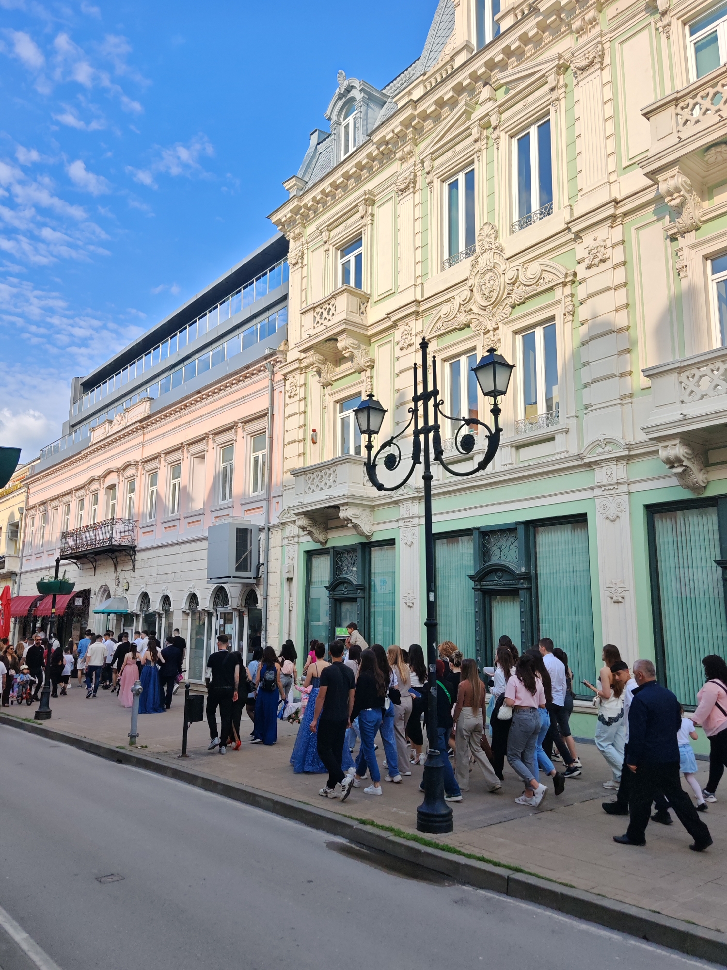 The image depicts a street scene in Ruse, Bulgaria. A large group of people is walking along a sidewalk adjacent to two distinct buildings. One building is a classic European structure with ornate detailing, balconies, and a cream and pale green color scheme. Next to it is a more modern building with a glass facade. A vintage-style street lamp stands on the sidewalk. Most people appear to be young adults, some dressed in what looks like formal wear, possibly for an event. The sky is blue with scattered clouds, suggesting it is daytime.