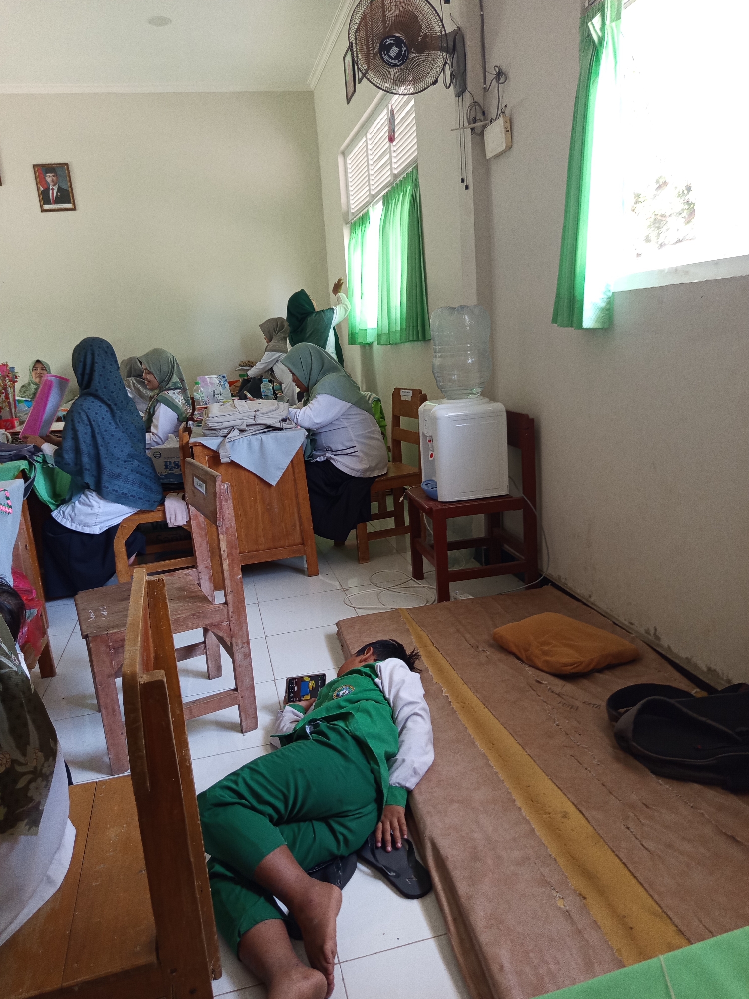 In a classroom in Lumutan, Indonesia, several young female students are seated at desks, some wearing hijabs. One student in the foreground is lying on the floor asleep in a green uniform. A water cooler is visible, along with various classroom items such as books, chairs, and a fan.