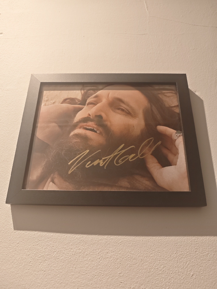 A framed portrait hangs on a light-colored, textured wall indoors. The frame is dark, appearing black or dark grey, and casts a shadow on the wall below and to its left.
The photograph within the frame is a close-up, sepia-toned or desaturated portrait of a man. He has a full beard and long dark hair, and his head is tilted back, gazing upwards with an intense or emotional expression; his mouth is slightly open. A hand, possibly his own, with a distinct ring on one finger, is visible near his temple, seemingly supporting his head.
Prominently displayed across the lower portion of the man's chest and neck in the photograph is a golden, handwritten signature. The signature clearly reads "Vint Hall".
The wall shows minor textural imperfections and subtle undulations. While the provided context suggests London, United Kingdom, there are no specific visual cues within the image itself to confirm this location. The lighting suggests an indoor environment.