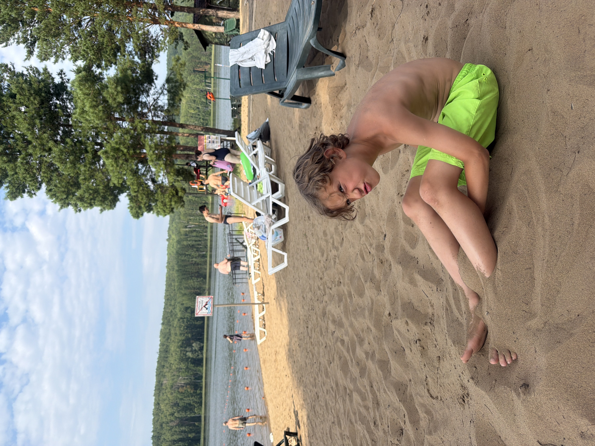 The image depicts a sandy beach on a sunny day, likely in Chaykovsky, Russia. In the foreground, a young boy with curly hair is partially buried in the sand, wearing bright green swim trunks. He is leaning forward, looking towards the camera with a playful expression. Behind him, several lounge chairs are scattered on the sand, some occupied by people. One lounge chair has a towel draped over it. Further back, a body of water, likely a lake or river, is visible, with trees lining the opposite shore. Several people are in the water, some standing and others appearing to swim. Orange buoys are floating in the water, possibly marking a swimming area. A sign with text and a graphic is partially visible near the water's edge. The overall scene suggests a relaxed summer day at a beach resort, with people enjoying outdoor recreation.