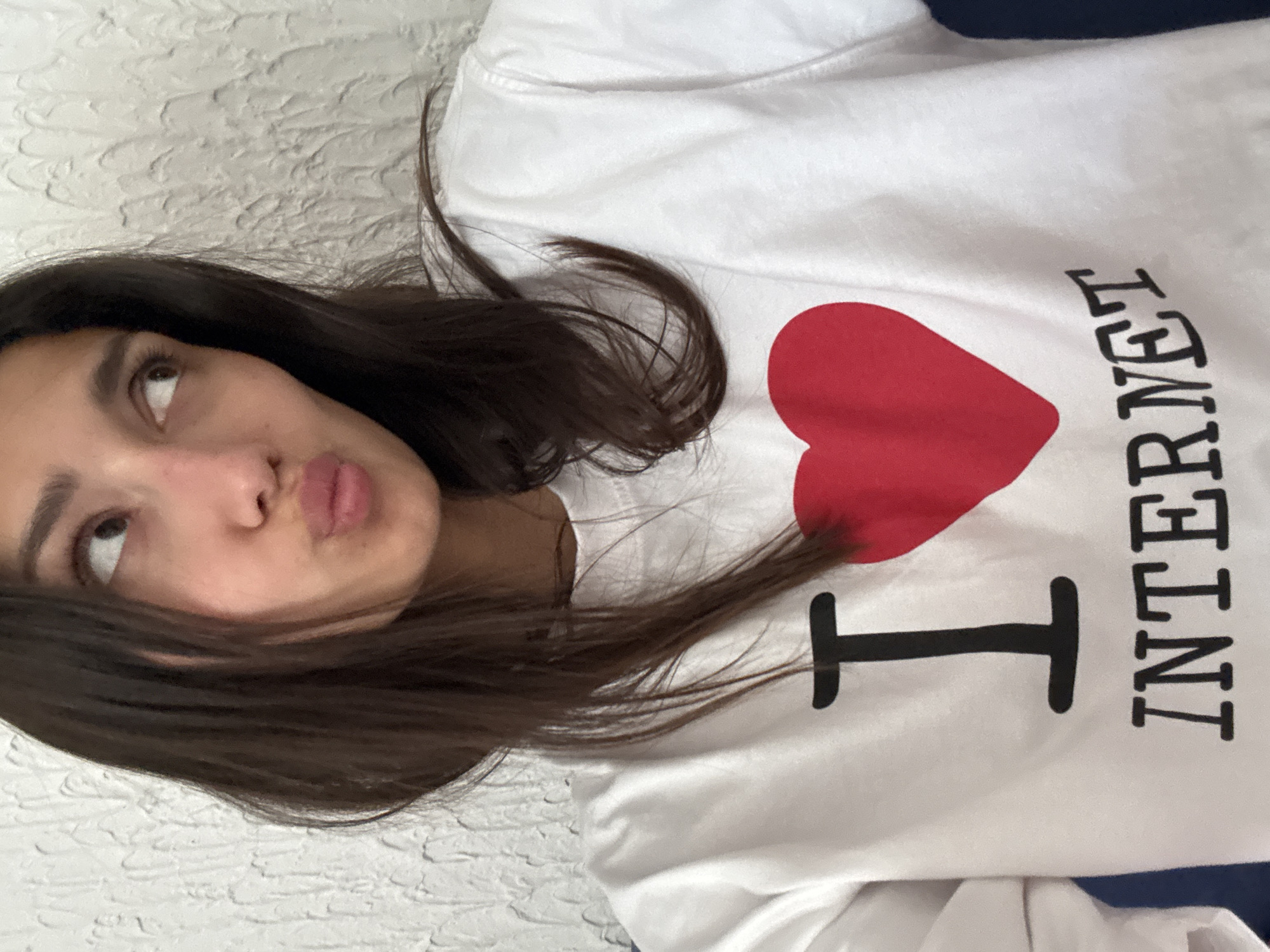 A close-up, angled shot captures a young person, likely female, from the chest up. She has long, dark brown hair that falls across her shoulder and the front of her white long-sleeved t-shirt. Her head is tilted, and she is looking upwards and slightly to the right, with a pouting expression on her lips. Her eyes are light brown.
She is wearing a white t-shirt that prominently features text and a large red heart. The design reads "I (red heart) INTERNET". The "I" and "INTERNET" are in black, bold, sans-serif letters, with "INTERNET" oriented vertically along the side of the shirt.
The background appears to be a light-colored, textured wall or surface, possibly stucco or a similar finish, partially visible above and below her head. The overall impression is a casual, indoor setting, possibly a selfie given the angle and proximity. The location context provided for the image is Zankovići, Montenegro.