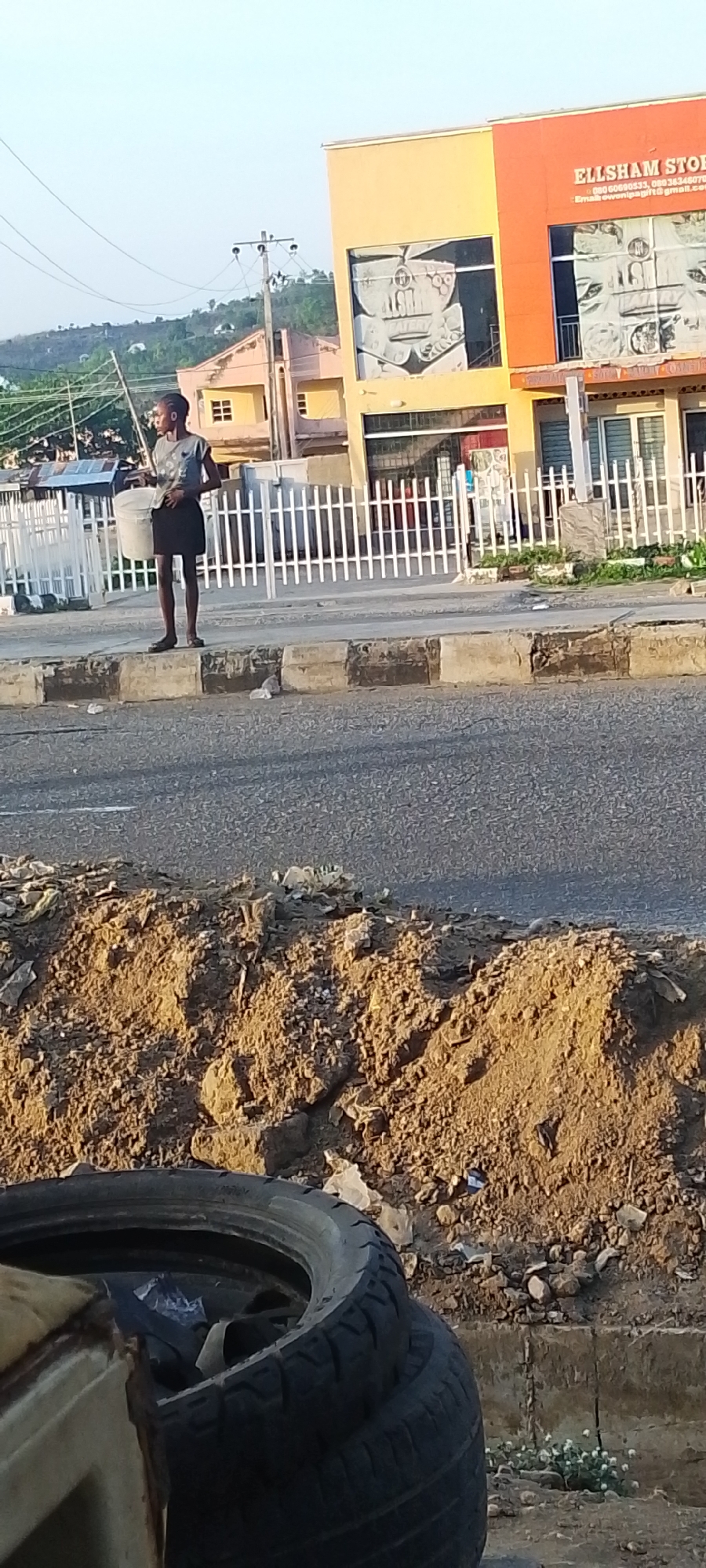 A young person stands on a curb on a city street in Lokoja, Nigeria, holding a pail. Across the street behind a white picket fence is a building with the words "ELLSHAM STORES" on the upper right side. The building is painted yellow and orange and has a black and white mural on its facade. The street in the foreground has a pile of dirt on the side with tires and trash. A hill or mountain is visible in the background.