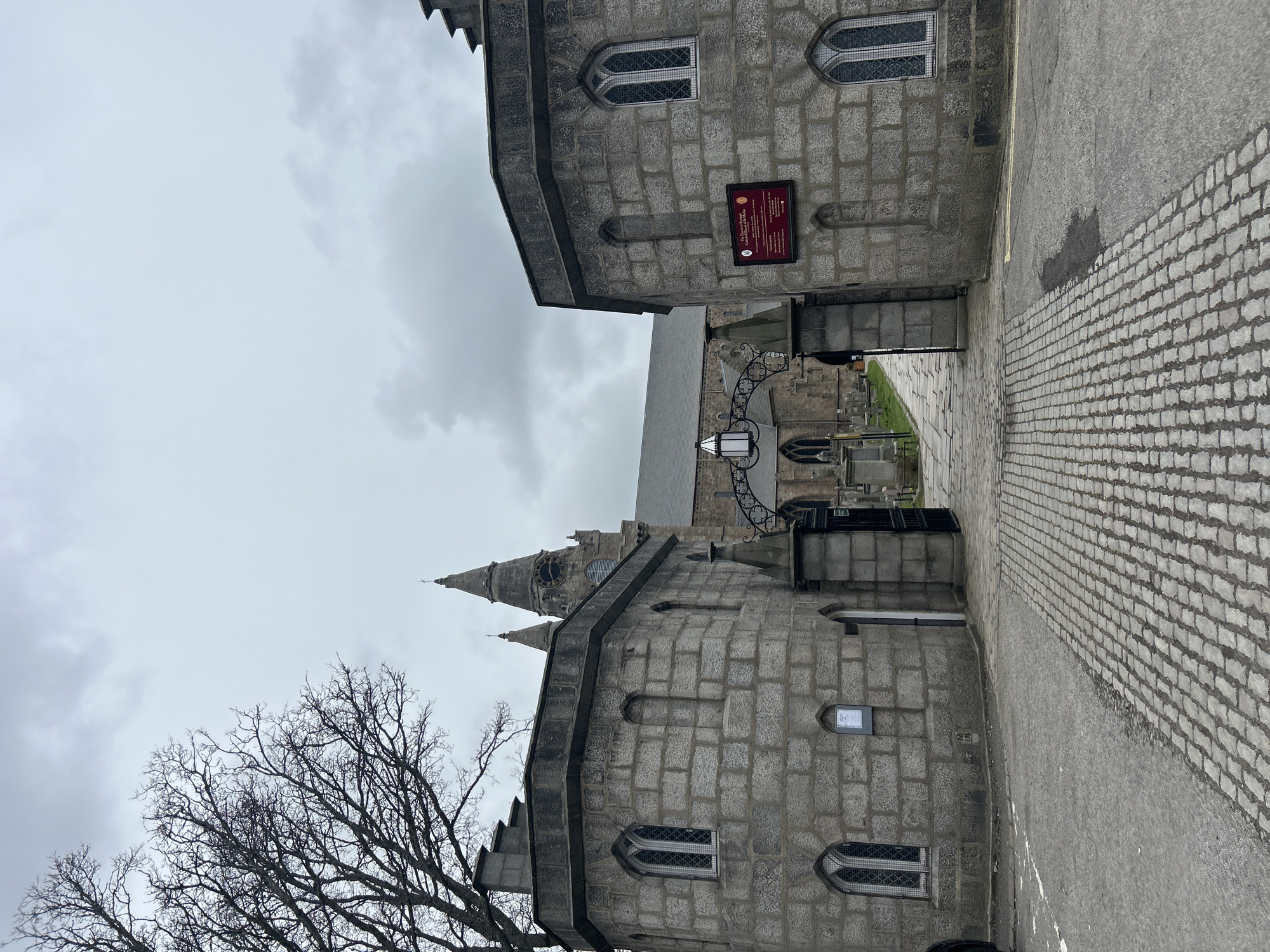 The image depicts a view in Aberdeen City, United Kingdom, showing two stone buildings lining a narrow walkway or street. The buildings are constructed from grey stone blocks, and feature arched windows with lattice-style panes. A sign is attached to the wall of one building. The walkway leads to a church with a steeple in the background. A wrought-iron gate and lamppost are visible along the walkway. A bare tree branch is visible in the lower left corner. The sky is overcast. The walkway is paved with light-colored rectangular stones.