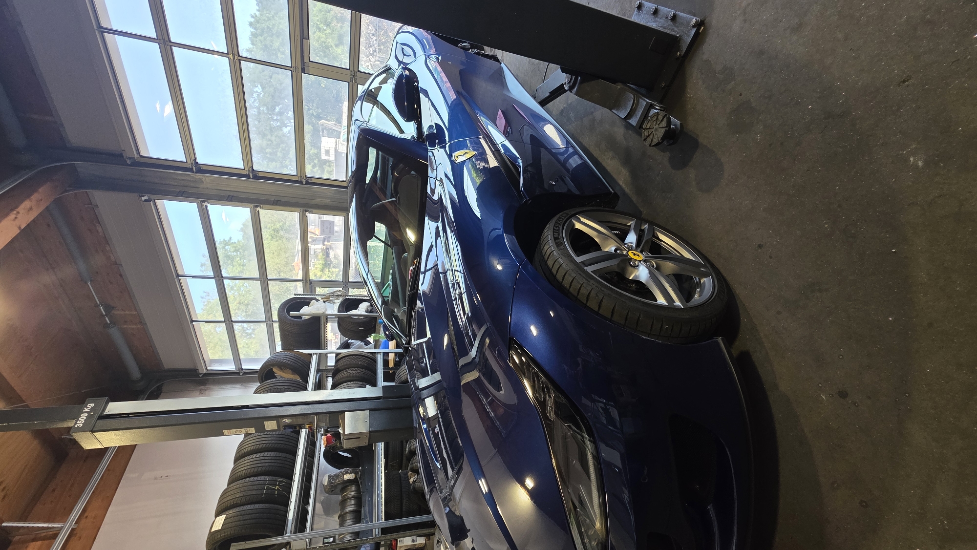A blue Ferrari is parked inside a garage. The car is on a hydraulic lift. There are shelves of tires in the background, and large glass doors with a view of trees and the sky. The garage has a wooden ceiling and concrete floor. A person is visible in the driver's seat of the Ferrari.