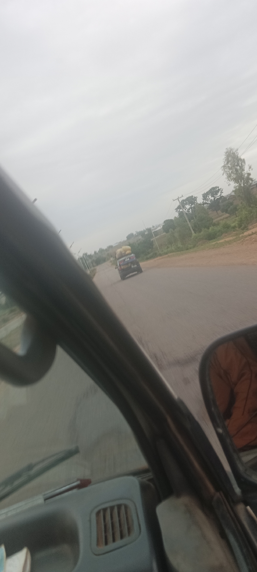 The image shows a road in the city of Barakin Kuru III, Nigeria. The photo was taken from inside a moving vehicle. Another car, with items piled on its roof, is visible further down the road. The sky is overcast. The side view mirror is visible. Trees line the roadside.