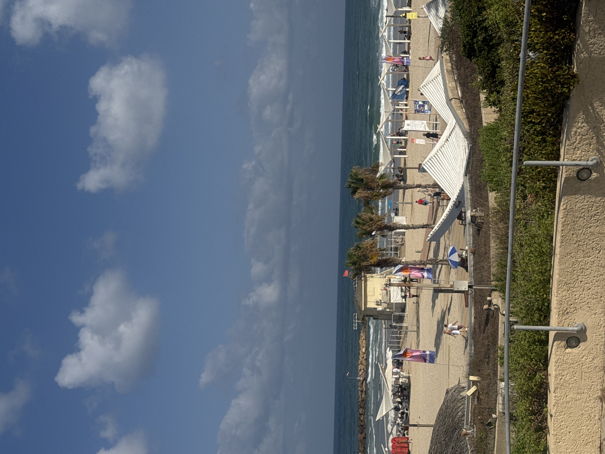 This image captures a lively beach scene in Herzliya, Israel, under a bright, partly cloudy sky. The foreground features a textured, light-colored wall with a metal railing, behind which lush green bushes are visible. A pathway slopes down towards the beach.
The beach itself is a sandy expanse dotted with numerous white sunshades. People are scattered across the sand, some relaxing under the shades, others walking or engaged in activities. A few palm trees stand tall, adding to the tropical atmosphere. Flags, one blue and white and two pink, are planted in the sand, adding splashes of color.
In the middle ground, a yellow lifeguard station stands prominently. A breakwater made of large rocks extends into the turquoise sea, where gentle waves are breaking. The sea stretches to the horizon, meeting the sky. The sky is a vibrant blue, with scattered white cumulus clouds. The overall impression is one of a pleasant, sunny day at a popular Israeli beach.