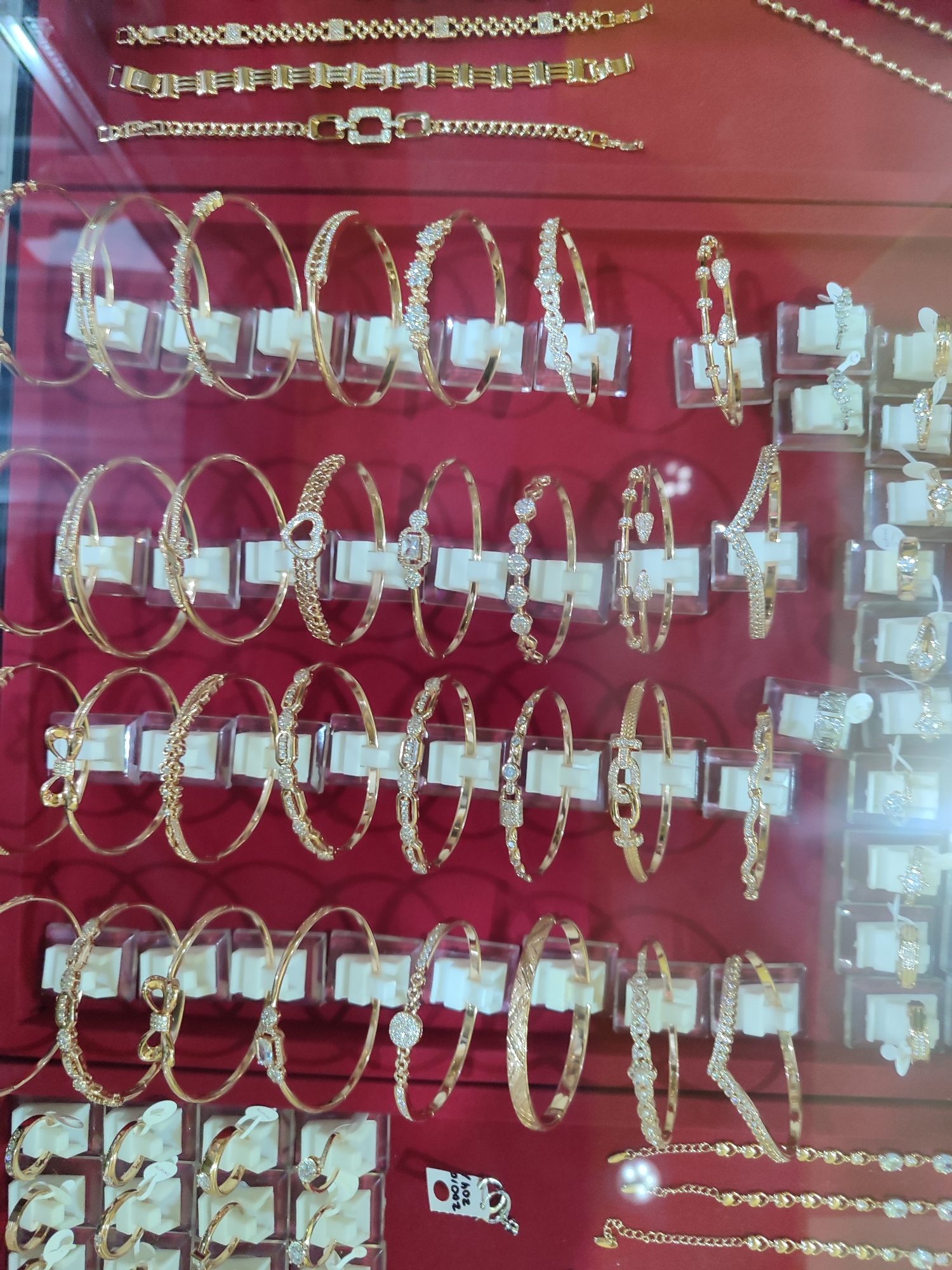 The image displays a close-up, eye-level view of a jewelry store display case in Randudongkal, Indonesia. The case is filled with an assortment of gold-colored bracelets and necklaces, many adorned with small, clear stones, likely diamonds or cubic zirconia. The jewelry is neatly arranged on white stands against a red background. In the bottom right corner, a tag with the numbers "20010 204" is visible, suggesting inventory or pricing information. No people are visible in the image.