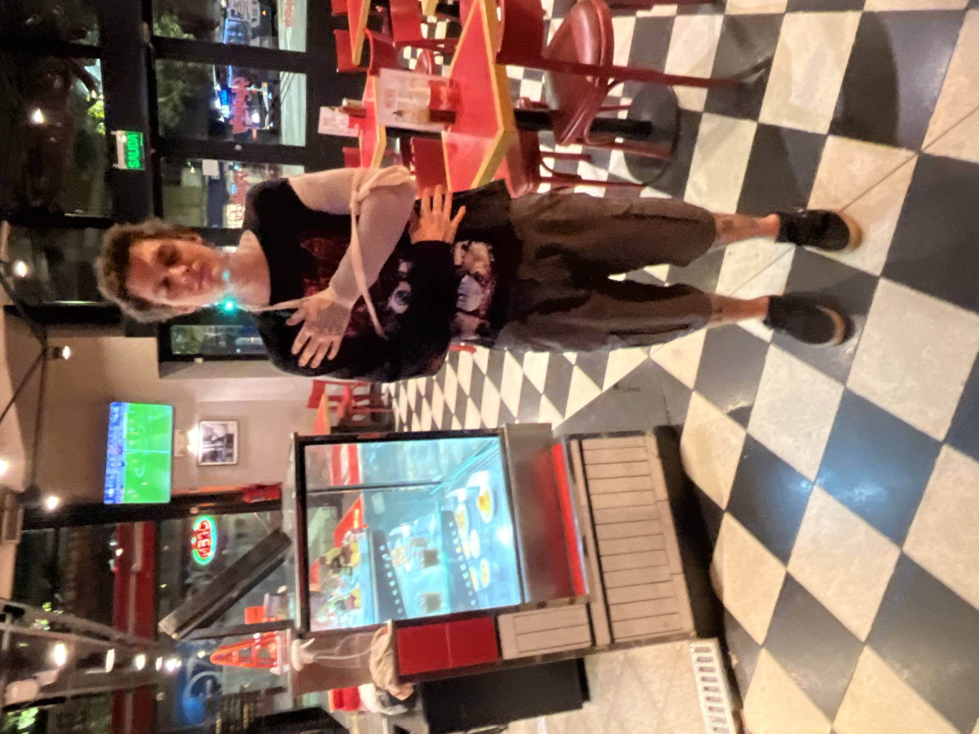 The image captures a young adult standing in a casual, indoor commercial setting, likely a diner or fast-food restaurant, during the evening. The subject, a person with dark, curly hair, is wearing a dark t-shirt with a graphic print that resembles a stylized face or skull, dark cargo pants, and dark sneakers with light soles. One arm is folded across their chest, and a light-colored wristband or bandage is visible on one wrist. They are looking directly at the camera with a slight smirk. A prominent green light reflects off their chest and neck area.
The environment features a distinct black and white checkered tile floor. Behind the person are red tables and chairs, characteristic of a diner, with some tables having star-shaped bases. To the left, a large refrigerated display case reflects interior lights and possibly merchandise. A flat-screen television is mounted on a wall in the background, displaying what appears to be a soccer match. Large windows line the back wall, showing reflections of outdoor lights and hinting at an urban street scene at night. A bright green sign with the Spanish word "SALIDA" (Exit) is clearly visible through one of the windows, indicating a Spanish-speaking location consistent with Buenos Aires, Argentina. The lighting is predominantly artificial, illuminating the scene brightly.