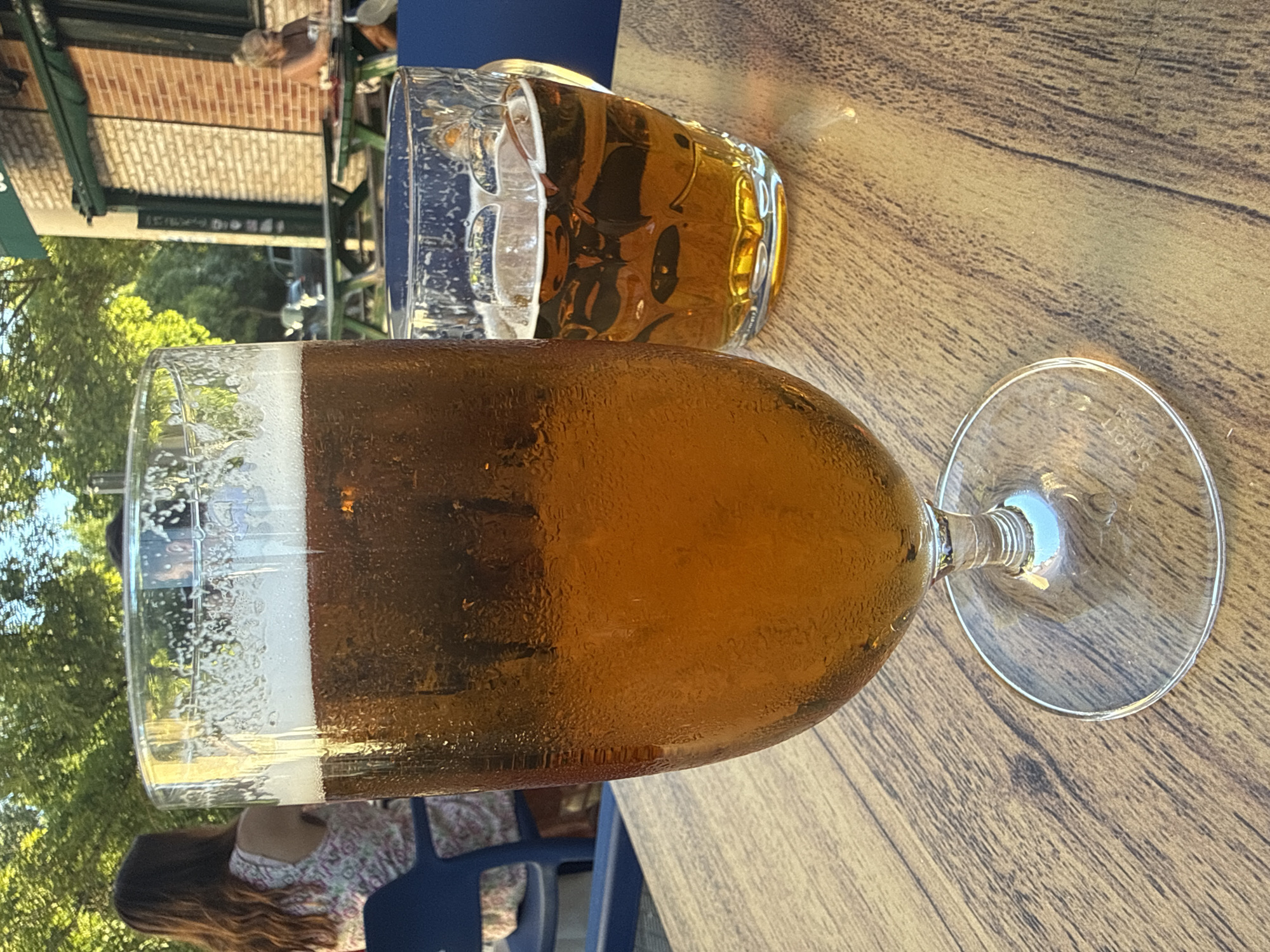 The image depicts an outdoor seating area at a cafe or bar in Pamplona, Spain. In the foreground, a large tulip-shaped glass filled with amber-colored beer and a frothy head sits on a wooden table. To the right, a smaller, handle-mug glass also contains beer. In the background, a person with long brown hair is seated at a table, wearing a floral-patterned dress. Further back, another person is visible at a table outside a building with a brick facade. Trees with green foliage and sunlight filter into the scene.