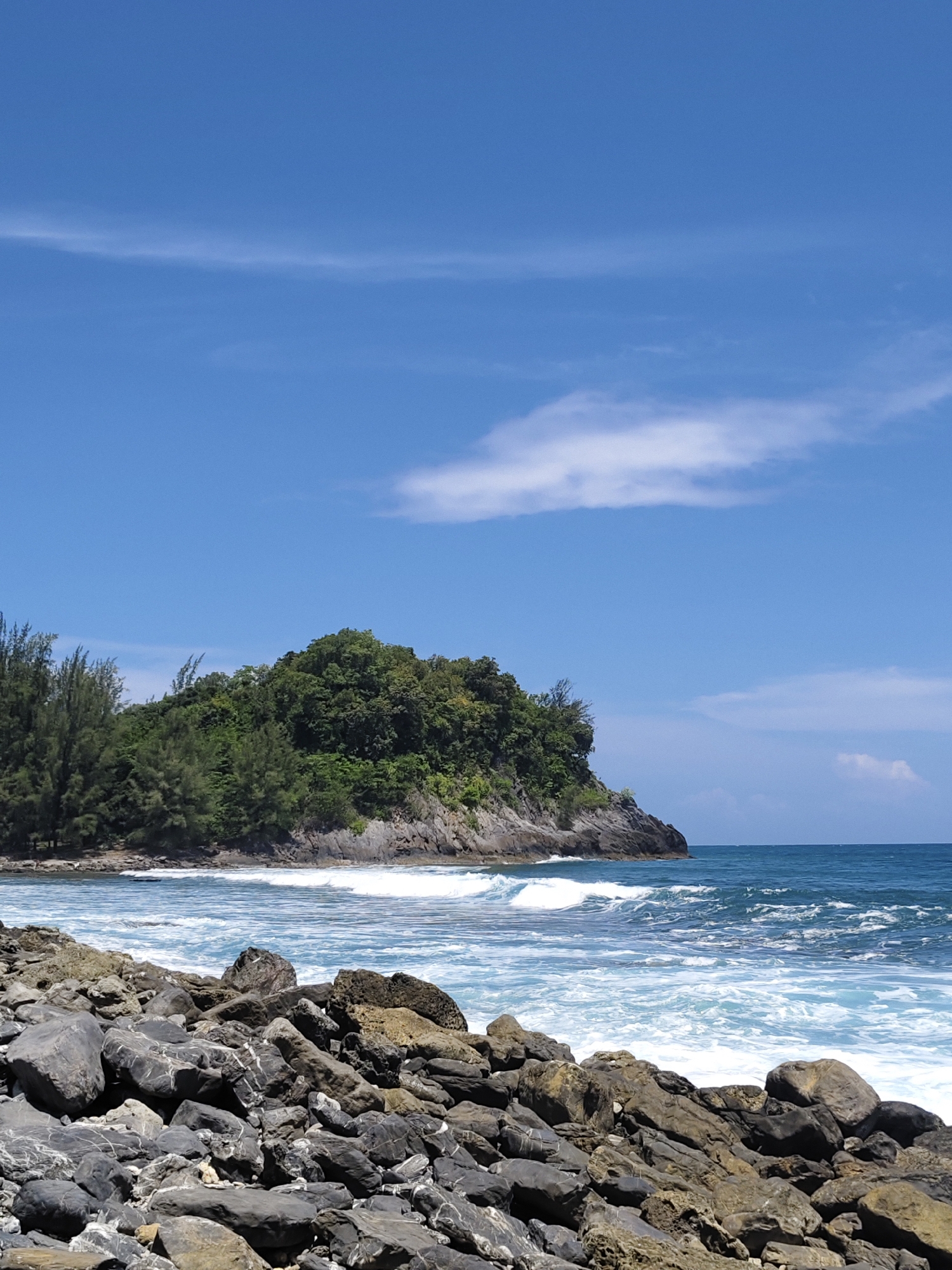 This image depicts a coastal scene in Naga Umbang, Indonesia. In the foreground, a rocky shoreline is strewn with various sizes of dark gray and brown stones. Beyond the rocks, the ocean presents a dynamic seascape with white-capped waves rolling towards the shore under a bright blue sky. A thin, wispy cloud stretches horizontally across the sky, with a puffier white cloud visible in the distance. To the left, a lush green, tree-covered hill juts out into the sea, forming a small cape. The vegetation on the hill is dense and appears to be a mix of trees. The water is a vibrant blue, suggesting a clear day. No people or animals are visible in the frame. The overall impression is one of a natural, unspoiled coastal landscape.