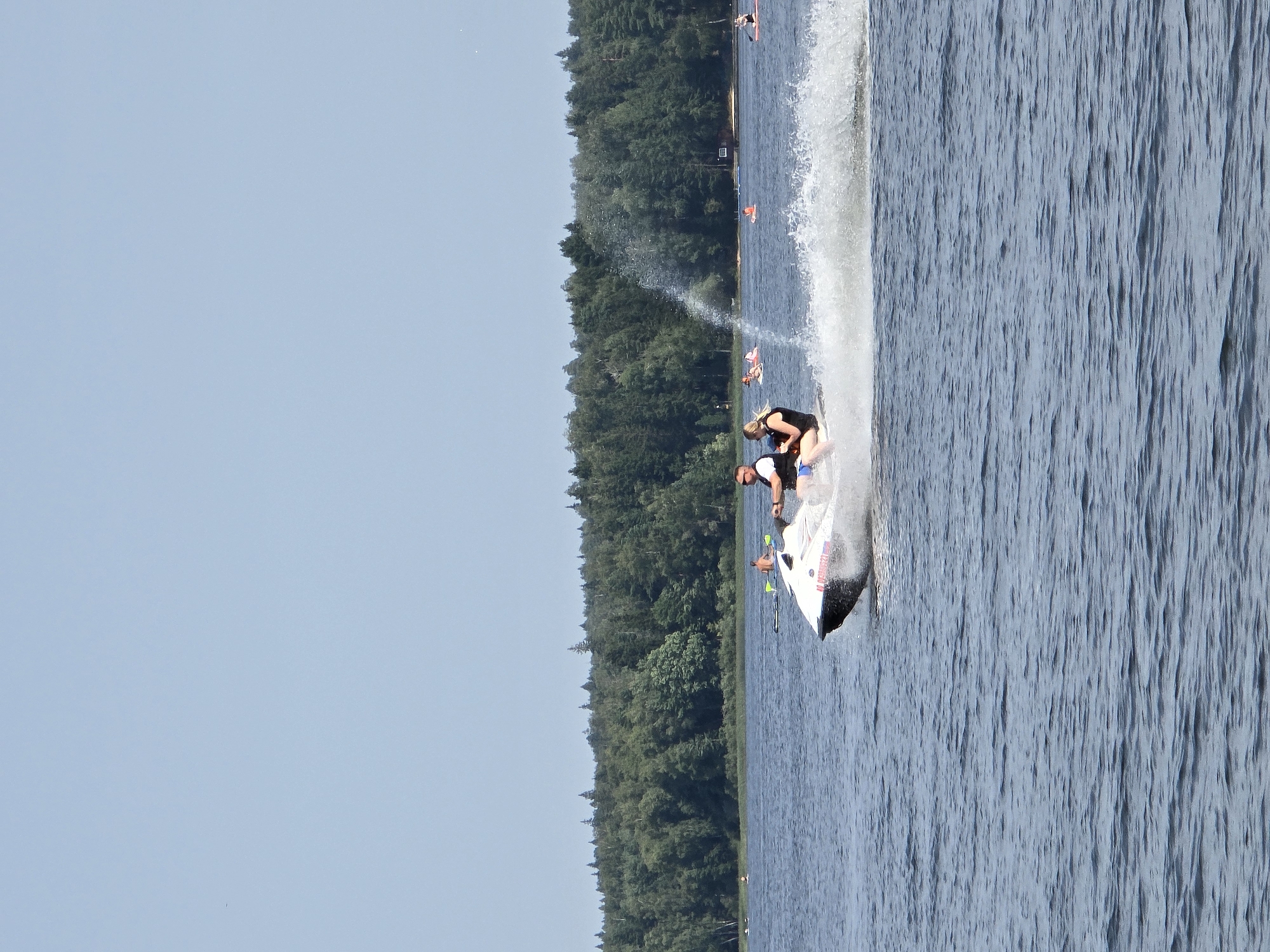 On a clear, sunny day, a white jet ski with two people on board is speeding across a body of water, creating a significant wake. The person in front, a male wearing sunglasses and a black vest, is operating the jet ski. The person behind him, a female also wearing a black vest, is holding on. In the background, a lush, tree-covered shoreline stretches across the horizon. Several other individuals are visible on the water, some on paddleboards and others swimming. The location appears to be a lake or a wide river. The image captures a recreational scene on the water, with a focus on the jet ski and its occupants. The general setting suggests a natural environment, possibly a park or a recreational area.