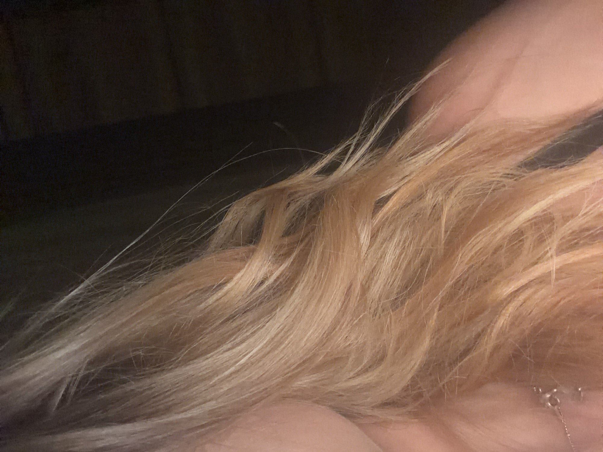 This is a close-up, indoor, waist-up shot of a person with light blonde, wavy hair, illuminated by warm lighting. The background is dark and out of focus, with a hint of what appears to be clothing visible at the bottom right. A delicate silver necklace is visible on the subject's chest, with a small pendant. There is no indication of the specific location within Enina, Bulgaria.
