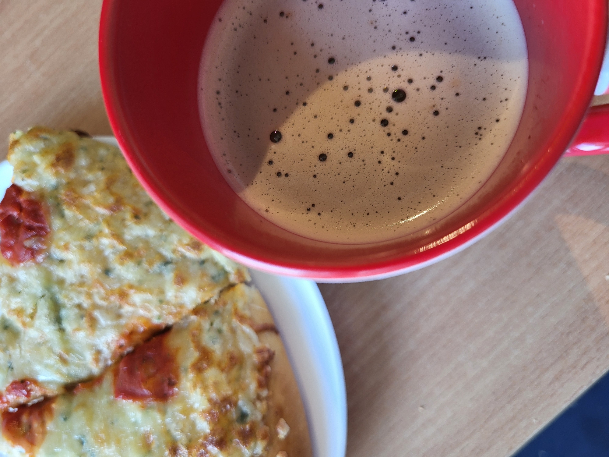 In an overhead, close-up shot, a slice of pizza rests on a white plate next to a red mug filled with coffee on a wooden table. The pizza appears to be thin crust with melted cheese and red sauce. The coffee has a frothy layer on top with small dark bubbles. The lighting in the image is soft, casting a gentle shadow on the coffee. The background is out of focus, but the wooden texture of the table is visible.