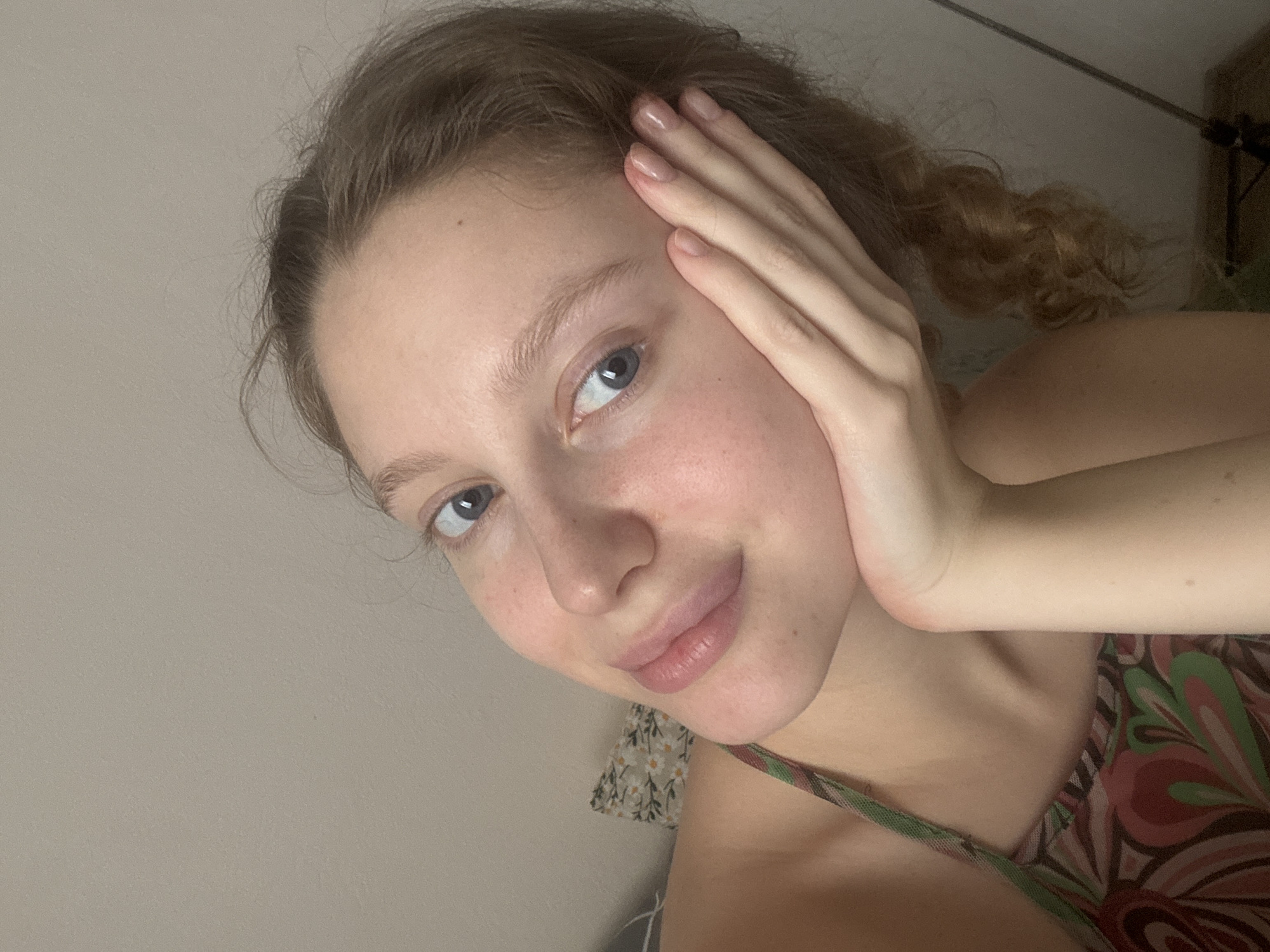 This is a close-up, overhead, selfie-style photo of a young woman with light skin, light brown hair in a braid, and blue eyes. She is wearing a camisole with a floral pattern of pinks, greens, and browns. Her right hand is resting on her face, near her temple. The background is a plain, light-colored wall, suggesting an indoor setting. The lighting appears to be soft, and there are no visible windows or indications of the time of day or weather. Her expression is relaxed, with a slight smile. The photo is taken from a slightly angled perspective. There is no visible text in the image.