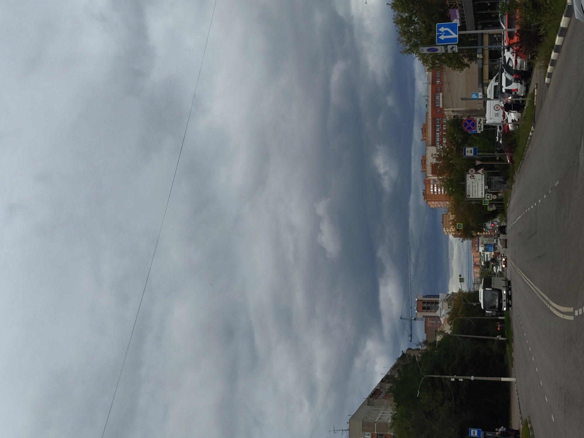 The image depicts a cloudy day in the city of Zhukovsky, Russia, likely on a wide avenue. The foreground shows a road with double white lines, indicating multiple lanes of traffic. A white bus is driving in the left lane, with other vehicles, including cars and what appears to be a van, visible further down the road.
On the left side of the road, there is a sidewalk and trees, with a bus stop sign. Behind the trees, a multi-story residential building with balconies is visible. Further in the background, a construction crane and unfinished buildings suggest ongoing development.
On the right side of the road, there are buildings with shops or businesses on the ground floor. Several cars are parked along the curb. Various street signs are present, including traffic signs for parking, traffic flow, and pedestrian crossings.
The sky is heavily overcast with dark, dramatic clouds, suggesting an impending change in weather. The overall scene conveys a typical urban environment under a moody sky.