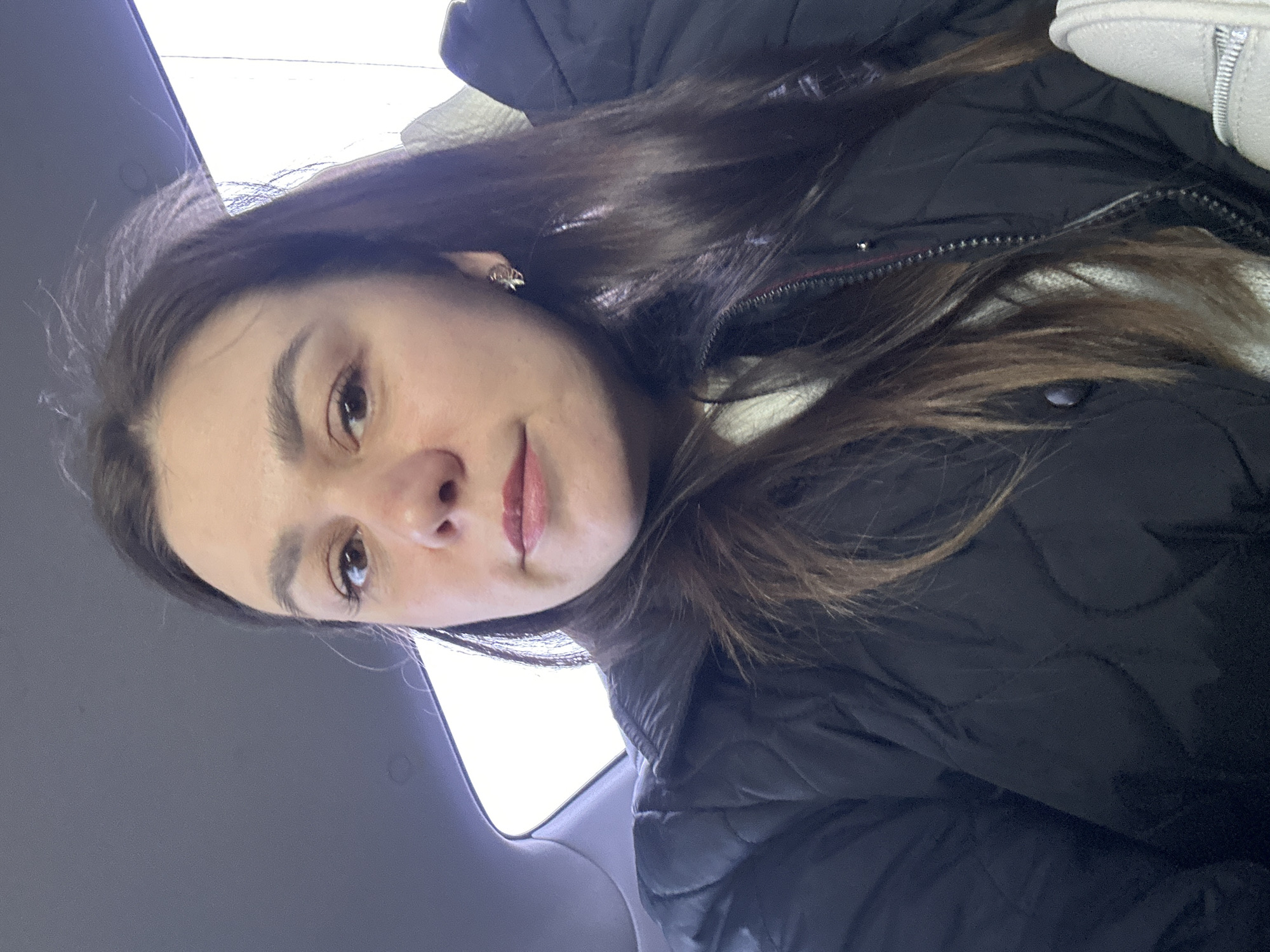 A woman is seated in a car. She is wearing a black, quilted jacket and a light-colored top. Her hair is long and dark. The car's interior is grey. Outside the window appears to be a bright, overcast day. The location is not discernible from the image to determine if it is in Chaikovsky, Russia.