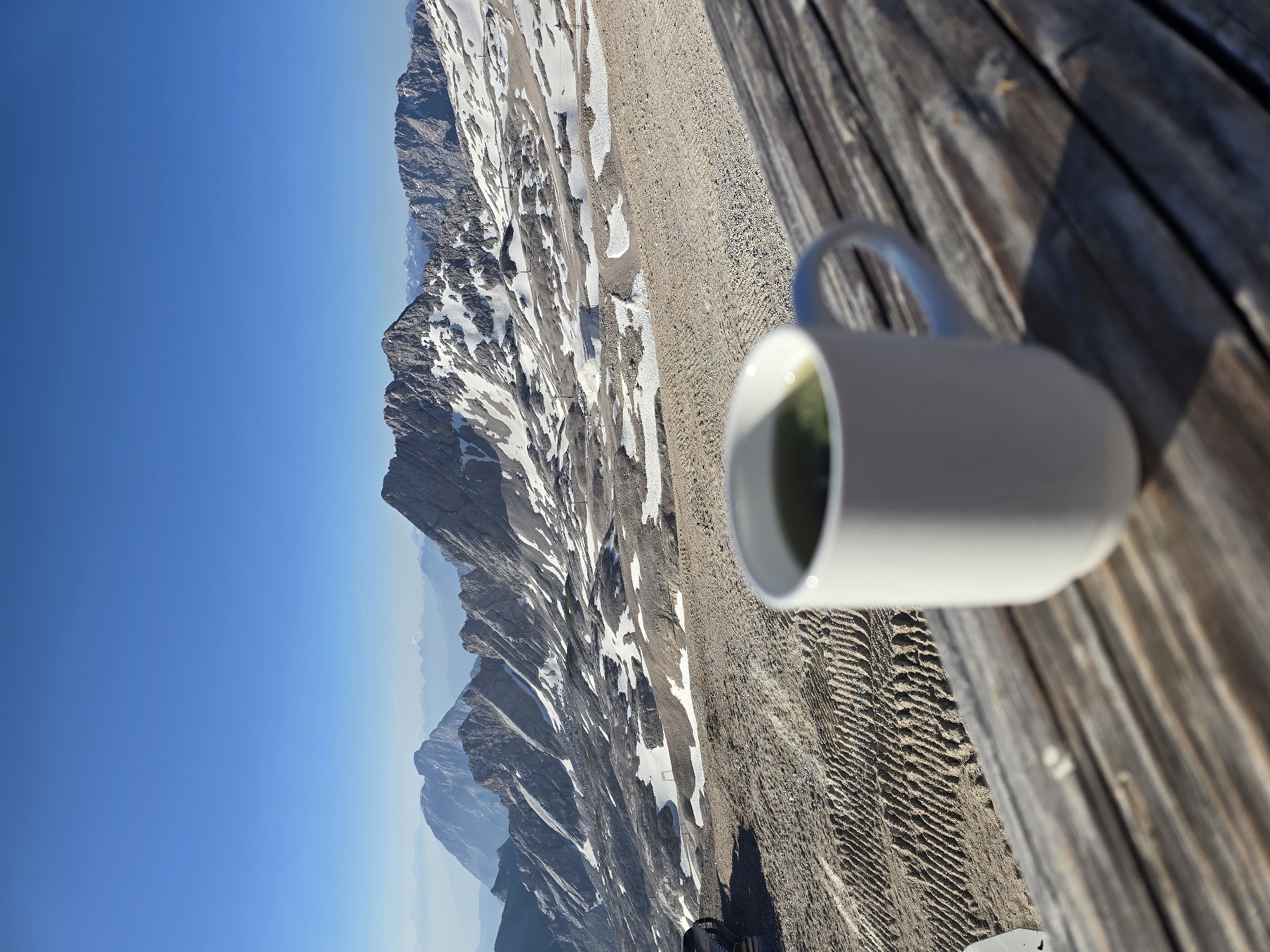 A white mug filled with a green beverage sits on a rustic wooden table, its surface weathered and sun-drenched. The mug is positioned in the foreground, slightly out of focus, with a pristine white exterior and a handle curved invitingly. Beyond the table, a breathtaking panoramic view of the Alps unfolds under a clear, deep blue sky. The mountains are rugged and majestic, with snow-capped peaks and rocky outcrops scattered across their slopes. The landscape appears vast and untamed, with evidence of human activity in the form of what appears to be a track or path carved into the textured, gravelly ground. In the far distance, layers of hazy mountains recede into the horizon. The scene suggests a serene moment of respite, with the warm sunlight casting shadows and highlighting the textures of both the natural and man-made elements. This view is characteristic of the Zugspitze, Germany's highest peak, located near Garmisch-Partenkirchen.