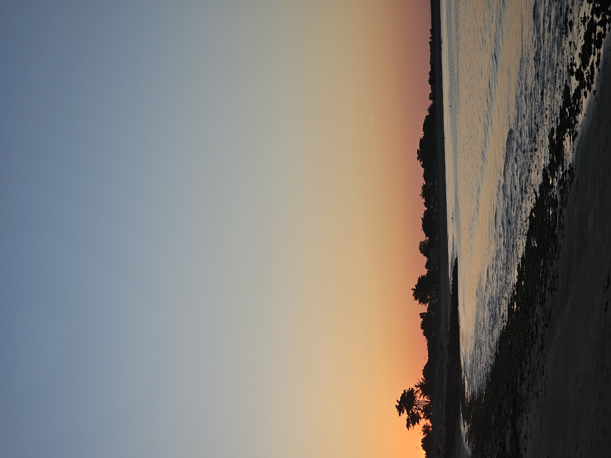 The image shows a beach scene at sunset in La Brée-les-Bains, France. The sky transitions from blue to orange. A dark treeline separates the sky from the beach and ocean. Waves are visible in the water, and the beach appears rocky. The image is taken at an angle.