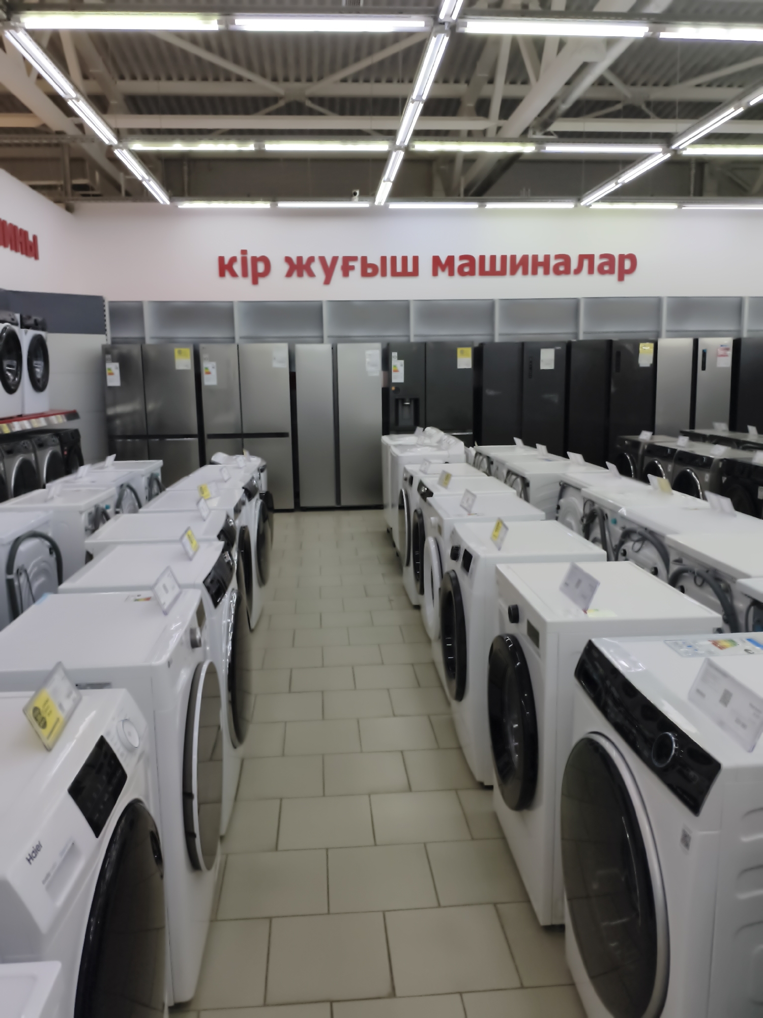 The image shows a retail store interior, likely a large appliance store, filled with numerous washing machines and refrigerators. In the background, a white wall displays red text that appears to be in Kazakh, with the phrase "кір жуғыш машиналар" visible, which translates to "washing machines." The foreground is dominated by rows of white front-loading washing machines, with their control panels and doors clearly visible. Some machines have yellow tags or labels. Further back, silver and black refrigerators are arranged in orderly rows. The store is well-lit by overhead fluorescent lighting, and the floor is tiled. The overall impression is one of a spacious retail environment dedicated to household appliances. Based on the prominent Kazakh text, this store is located in Aktobe, Kazakhstan. There are no people visible in the image.