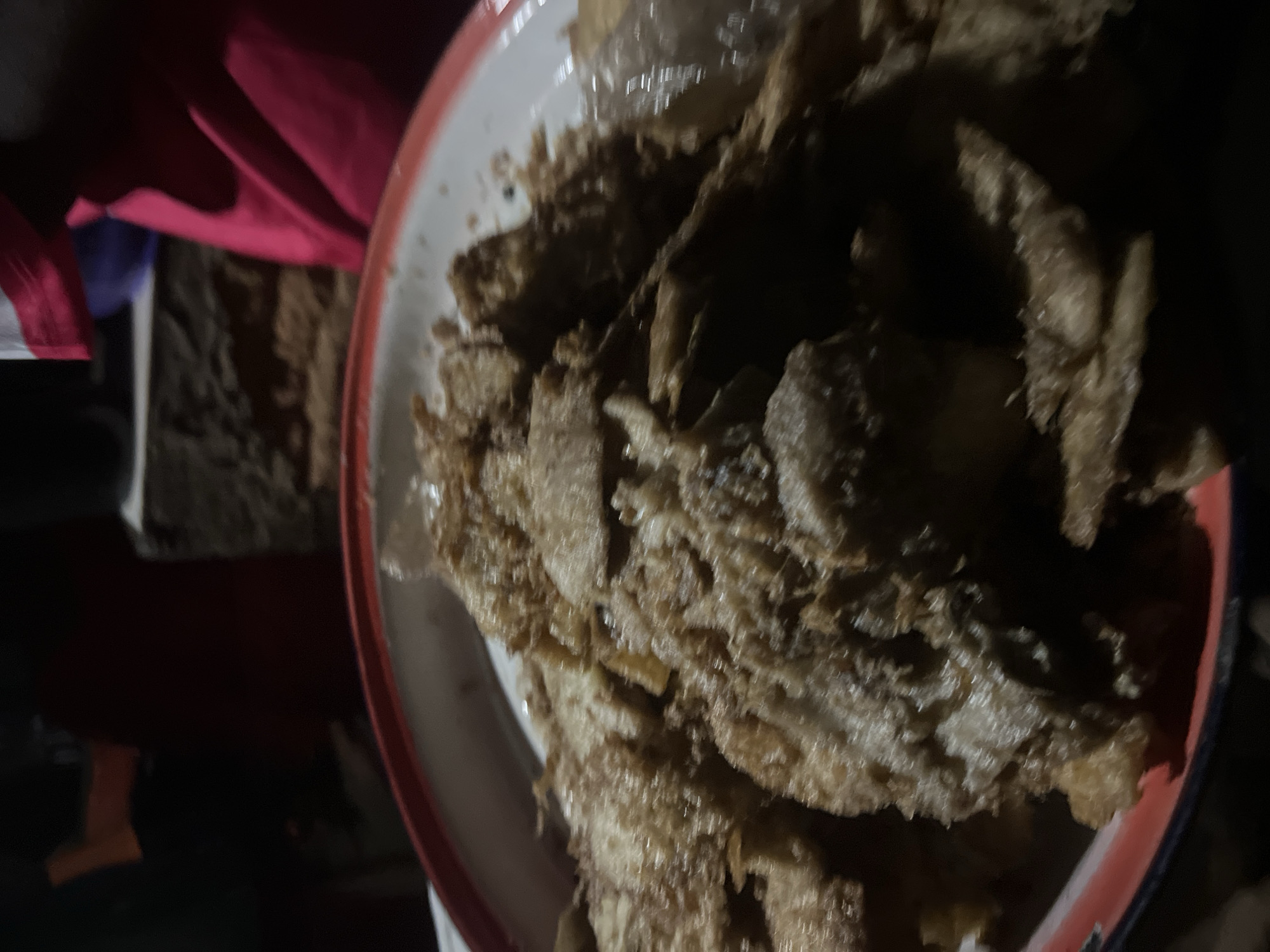 The image depicts a close-up view of a serving of fried plantains in a red-rimmed white plate. The plantains are sliced and appear to be deep-fried, with a crispy, golden-brown exterior. The dish is densely packed, with pieces overlapping and piled high. The lighting in the image is dim, suggesting it was taken at night or in a low-light environment. In the background, out of focus, are hints of a red cloth or garment and what appears to be a brick wall or textured surface. The overall scene is intimate and focused on the food. There is no visual information to pinpoint the location as Durumi, Nigeria.