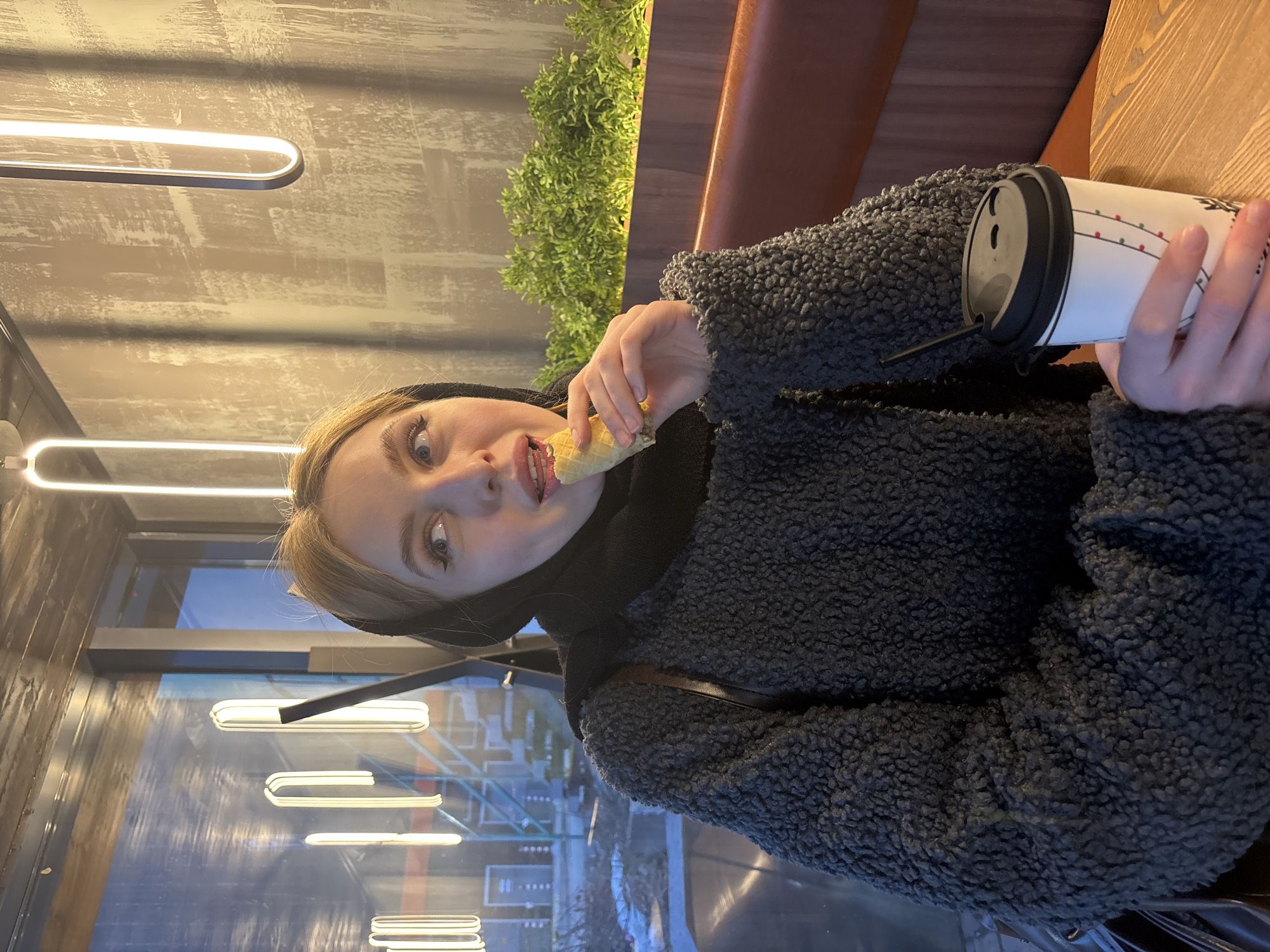 The image shows a young woman seated at a wooden table inside a modern cafe, consistent with a location in Perm, Russia. She has blonde hair partially covered by a black headscarf, wears a dark grey textured fleece jacket, and sits on a brown upholstered booth seat. Her right hand holds a light-colored rolled pastry, which she is bringing to her open mouth while looking upwards and to her left. Her left hand rests on a white disposable coffee cup with a black lid and straw, decorated with a festive light string design. In the background, a textured light-brown wall features multiple vertical, elongated oval LED light fixtures. To her right, a dark wooden planter box contains vibrant green foliage. Through a large window on her left, an exterior scene is visible with reflections of interior lighting and indistinct outdoor structures, possibly scaffolding or industrial elements, and patches of ground that appear covered with snow or debris under dim light.