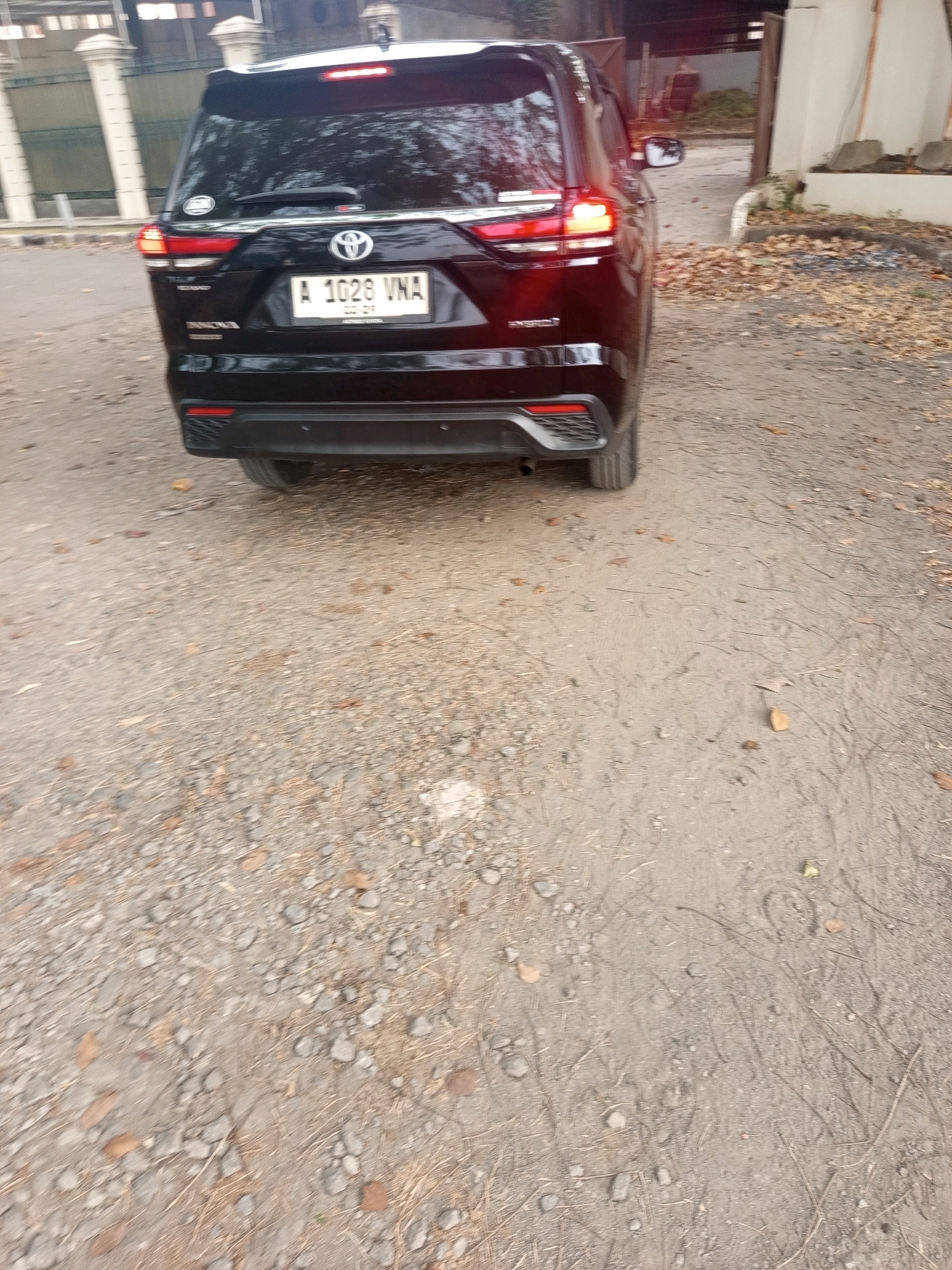 The image captures the rear view of a black Toyota Innova Hybrid parked on a dusty, gravelly surface, likely a street or driveway, in West Jakarta, Indonesia. The car's license plate reads "A 1028 VNA". Fallen leaves and small stones are scattered across the ground. In the background, a building with white pillars and a fence can be seen. The overall scene suggests an outdoor setting in an urban or semi-urban area.