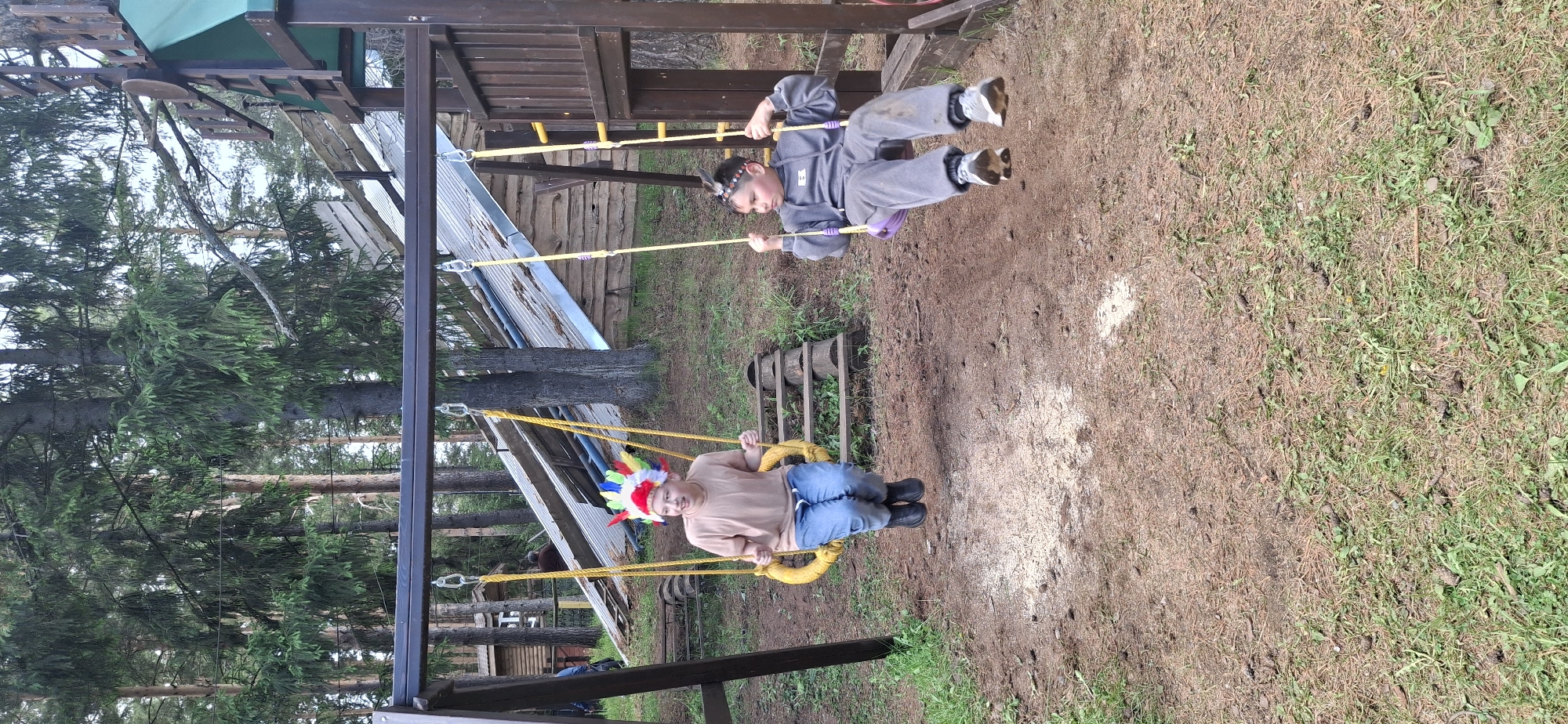 Two children swing on swings at a playground. One child wears a colorful feather headdress and blue jeans, while the other wears a gray tracksuit. The swings are part of a wooden playset, set against a backdrop of trees. The ground is a mix of dirt, wood chips, and grass. The image appears to be taken in Прикамский, Russia.