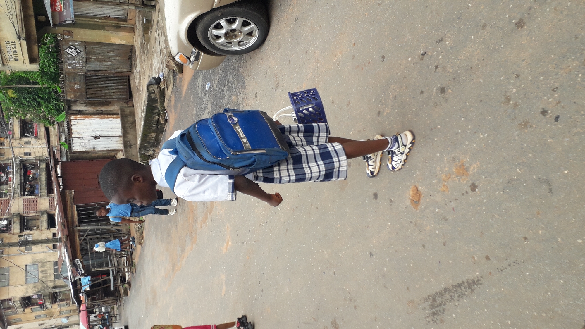 A young boy wearing a white shirt, blue and white plaid shorts, and blue and white sneakers is walking on a street in Onitsha, Nigeria. He is carrying a blue backpack and a blue plastic basket. He is facing away from the camera and to the right. To his left, a man wearing a blue shirt is looking at him. The background includes buildings, a car, and a street. A sign reading "THIS HOUSE IS NOT FOR SALE" is visible on one of the buildings. The street surface is a light grey color.