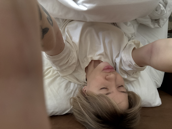 The image depicts a female individual in a selfie orientation, lying in a bed with light-colored bedding. Her eyes are closed, and her lips are pursed forward. She has short, light-colored hair, appearing somewhat disheveled. She is wearing a light-toned, short-sleeved t-shirt. Her head is positioned on a white pillow. Her right arm, extended towards the camera, shows a faint, raised linear scar on the inner forearm. A portion of her left arm is also extended, revealing a dark tattoo on the upper arm area. A small, silver stud earring is visible in her right ear. The background behind the pillow is dark, suggesting a headboard or wall. There is no visual information within the image to determine its specific location within Barcelona, Spain.
