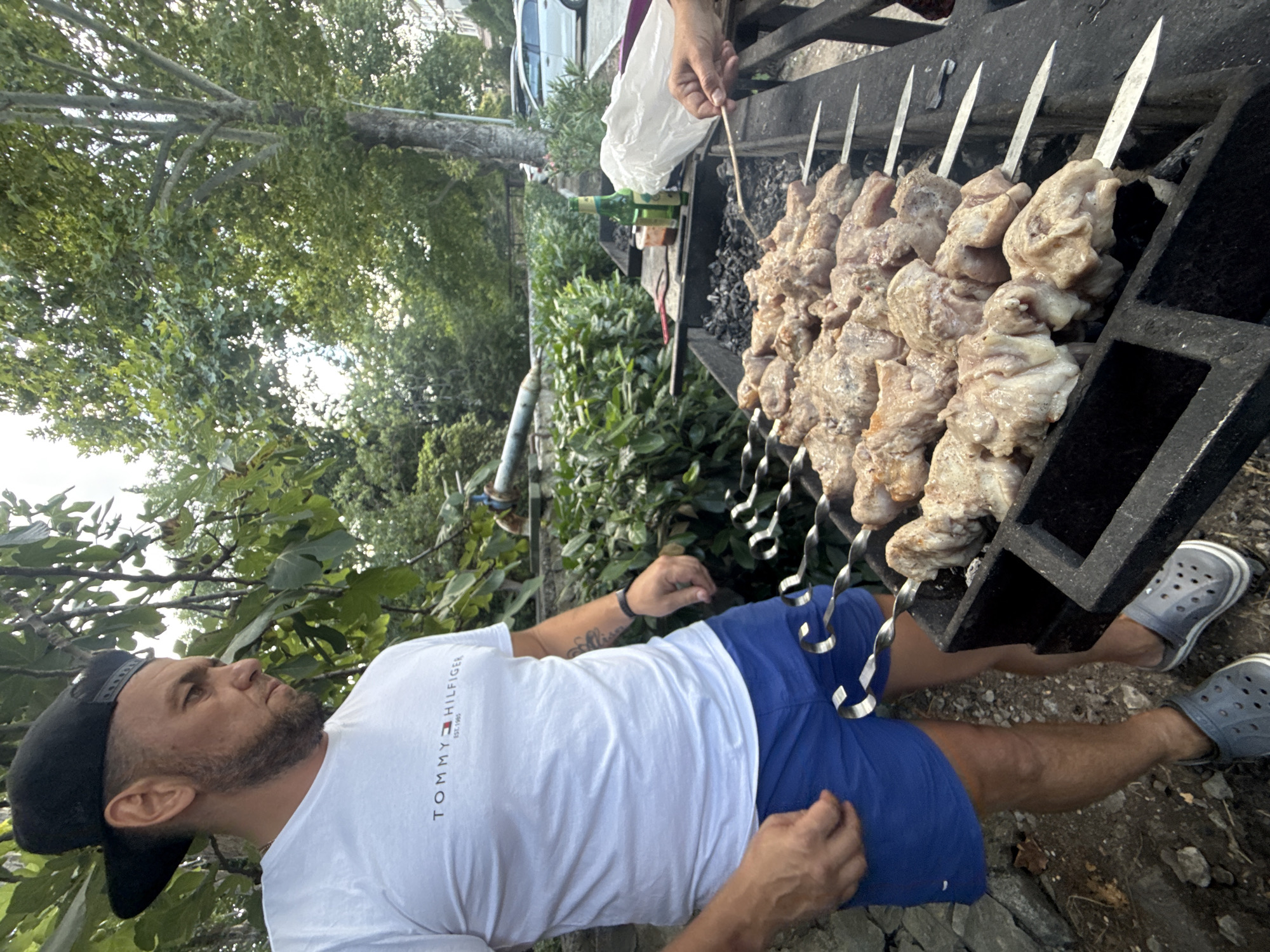 A man wearing a black baseball cap, white Tommy Hilfiger t-shirt, and blue shorts stands to the left of a grill laden with skewers of raw meat. He is looking down at the meat. Behind him, lush green foliage and trees suggest an outdoor setting, possibly a park or residential area. In the background, cars are visible parked along a street, indicating a semi-urban location. The grill contains several rows of skewers, each holding chunks of seasoned meat. A hand on the right side of the frame is holding a skewer, suggesting someone is actively engaged in preparing or tending to the barbecue. The overall scene depicts a casual outdoor gathering centered around grilling food.
