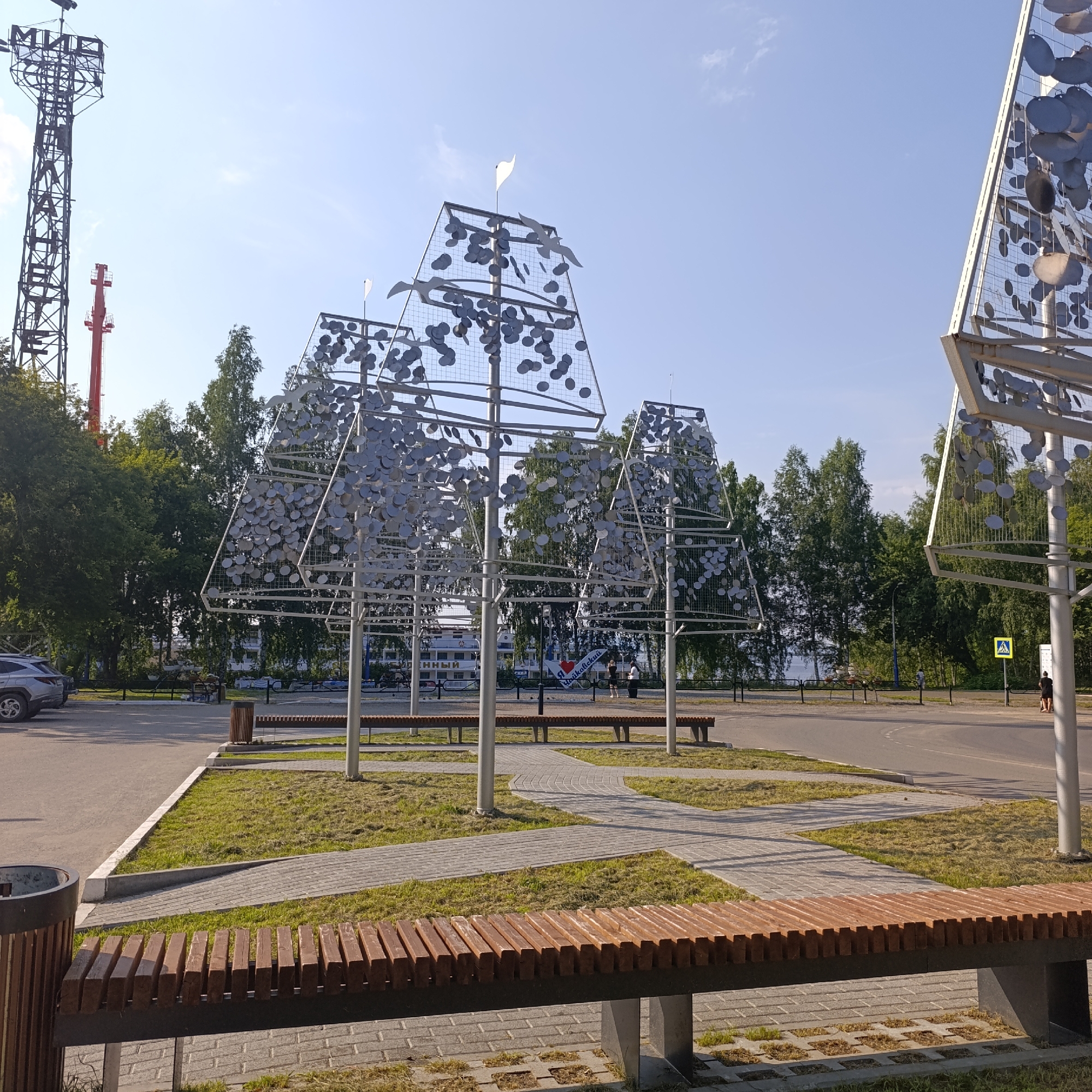 In Chaikovsky, Russia, an outdoor scene depicts a park-like area with modern sculptures. In the foreground, a wooden slatted bench faces a pathway paved with interlocking bricks. To the left, a cylindrical trash bin made of dark material and wood is visible. Behind the bench, a winding brick pathway leads through a grassy area with scattered patches of dry grass, interspersed with tall, slender metallic poles supporting abstract sculptures. These sculptures are pyramidal in shape, constructed from a metal frame covered in a mesh grid, adorned with numerous circular metallic discs that resemble leaves or petals. Several of these sculptures are arranged in a cluster. White flags are positioned atop some of the sculptures. In the background, trees line the horizon, with a body of water visible beyond them. To the far left, a tall, lattice-like tower and a red structure are prominent against a clear blue sky with a few scattered clouds. A parked silver SUV is seen to the left of the pathway. Further back, beyond the sculptures, a road is visible, with a pedestrian crossing sign and two individuals standing near it. Another bench is positioned in the mid-ground.