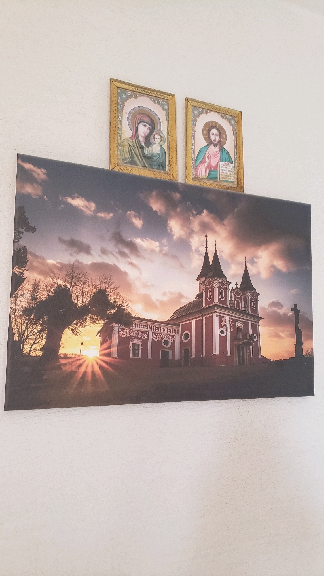 Two framed religious icons hang on a white wall. To the left is an icon of the Virgin Mary holding the infant Jesus. To the right is an icon of Jesus Christ. Below the icons is a large canvas print depicting a red church with spires at sunset. The sun's rays emanate from behind a silhouetted tree on the left side of the canvas. A crucifix stands to the right of the church. This church is likely the Church of the Nativity of the Virgin Mary in Prešov, Slovakia, known for its striking red exterior and baroque architecture.