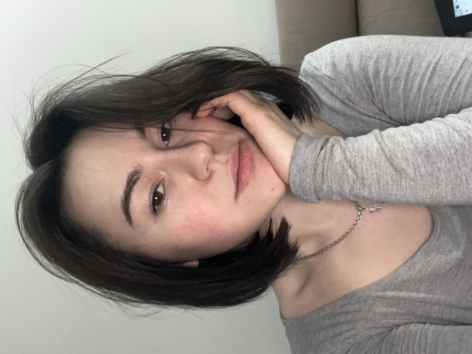 A young woman with short, dark hair and light skin is the focus. She's wearing a gray, long-sleeved shirt and a silver necklace. Her face is partially obscured by her hand, which rests on her cheek. She looks directly at the camera. The background is a blurred combination of white and beige tones, possibly a wall and a couch.