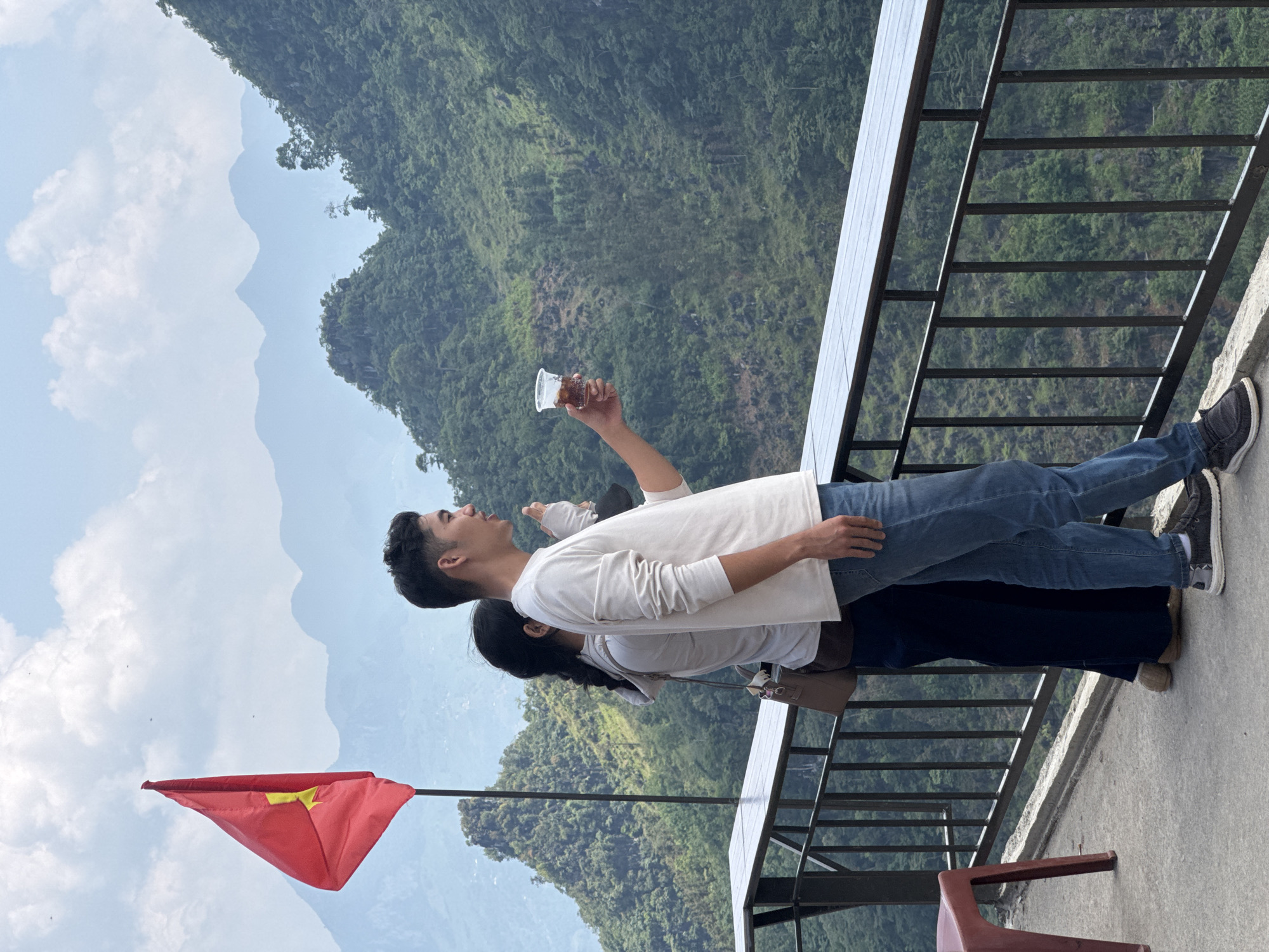 Two people stand on a platform overlooking a mountainous landscape in Lũng Phìn Commune, Vietnam. The man, wearing a long-sleeved white shirt and jeans, holds up a plastic cup with a dark liquid. The other person, partially obscured behind him, has long dark hair. A Vietnamese flag is positioned to the left of the scene. A metal railing borders the platform, and chairs are partially visible at the edge. The background features lush green mountains and a partly cloudy sky.