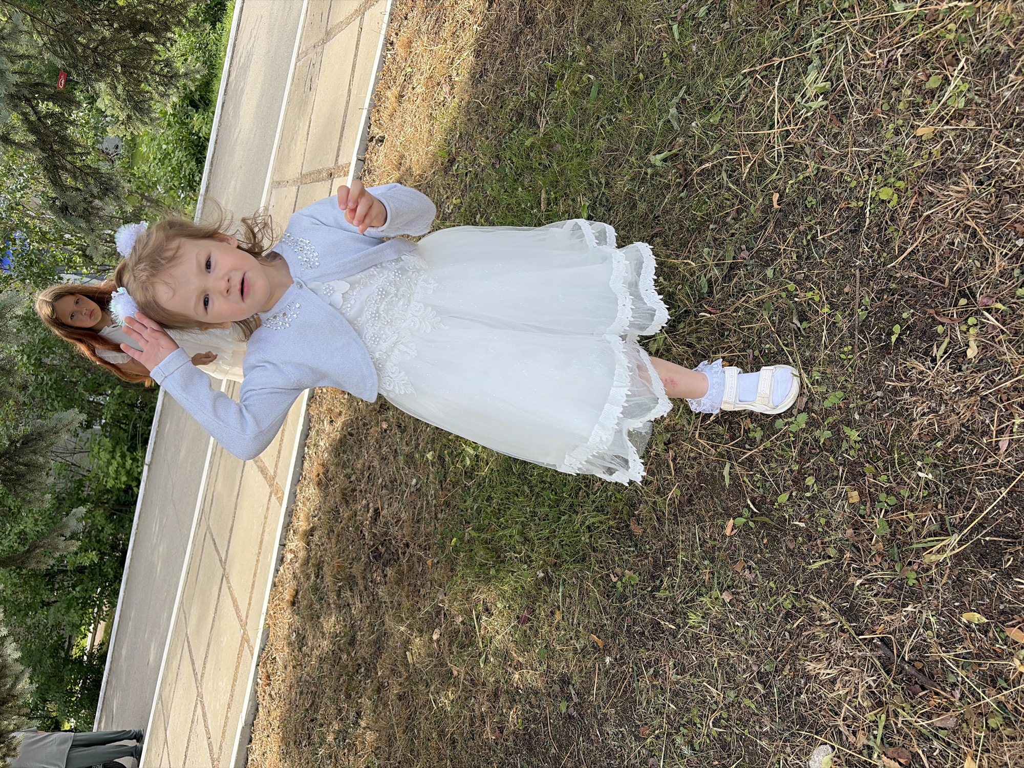 The image shows a young girl in a white dress and a light blue cardigan, standing on a grassy area next to a paved walkway. She has her right hand raised and appears to be looking at the camera. In the background, another girl in a white dress with long brown hair is visible. The environment suggests an outdoor setting, possibly a park or a street in Chaikovsky, Russia, with trees and foliage in the background. The girl is wearing white sandals with straps. The overall scene captures a candid moment, likely taken during an outdoor event or outing.
