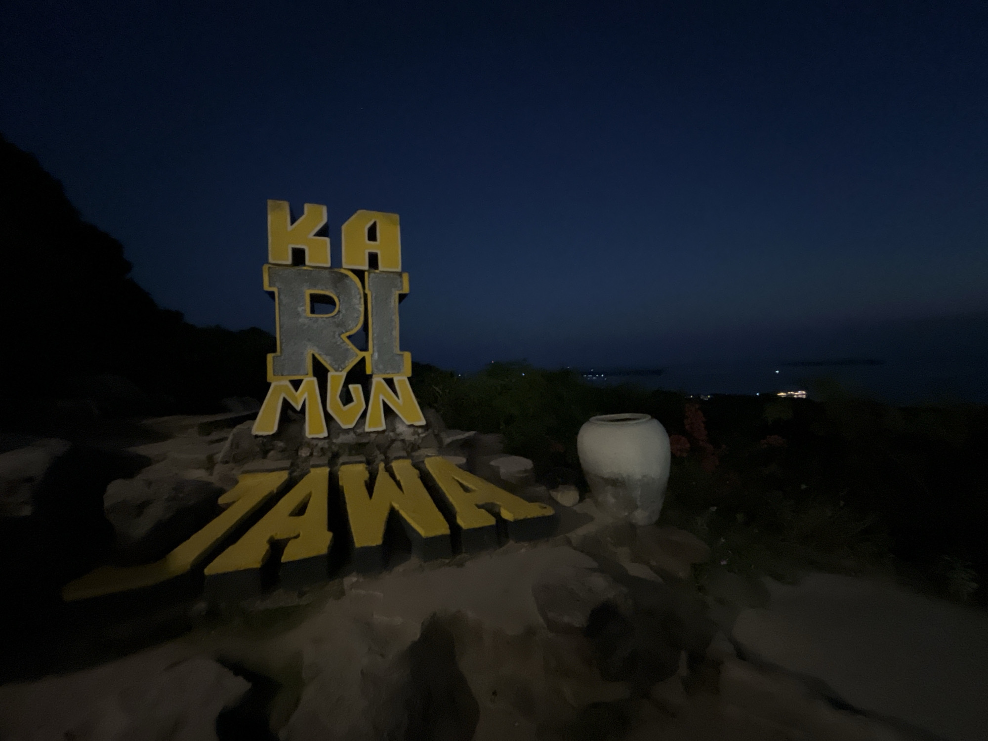 A large, three-dimensional sign in the foreground reads "KARIMUNJAWA." The letters are stylized and appear to be made of concrete with yellow detailing. The sign is set against a dark blue night sky, suggesting the image was taken after sunset. To the left of the sign, a dark, silhouetted hill or mountain slopes downward. To the right of the sign, a large, white ceramic pot sits on the ground. The surrounding area is dimly lit, with some vegetation and rocky terrain visible. In the distance, faint lights suggest a settlement or town, possibly on an island, with a body of water between the foreground and the distant lights. The overall mood is serene and peaceful, with a hint of mystery due to the low light conditions. The location is identified as Karimunjawa, Indonesia.