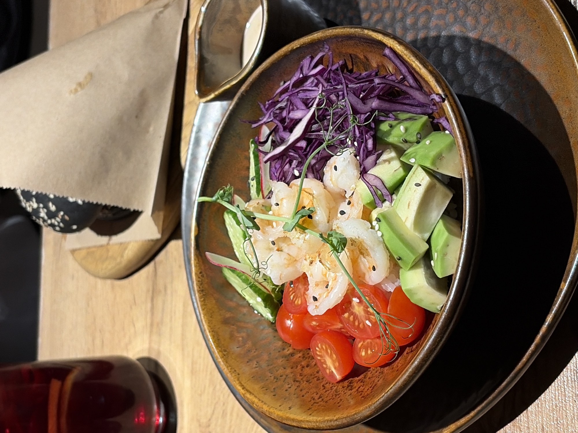 The image depicts a bowl of food, possibly a salad or poke bowl, with ingredients including shrimp, avocado, cherry tomatoes, shredded red cabbage, and greens. Sesame seeds are also visible. The bowl sits on a two-tiered plate. A brown paper wrapper, possibly for a sandwich, is partially visible in the upper left, along with a small bowl or container. A red object, potentially a bottle, is partially visible in the lower left. The surface beneath is a wooden tabletop. The location is indicated as Chaikovsky, Russia, although no specific landmarks or identifying features are present in the image to confirm this.