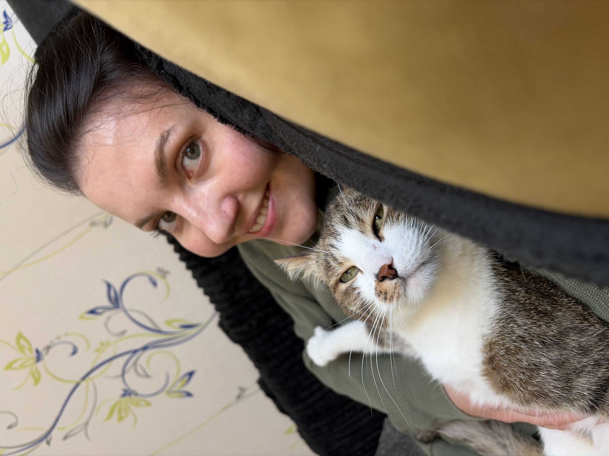 A woman with dark hair and green eyes is holding a tabby cat with white fur in her arms. The woman is smiling and looking at the camera. The cat is looking to the left of the camera with a neutral expression. They are positioned next to a black fabric covered object and a yellow object. The background includes a wall with a floral pattern in blue, green and grey. This photo was taken in the city of Chaikovsky, Russia.