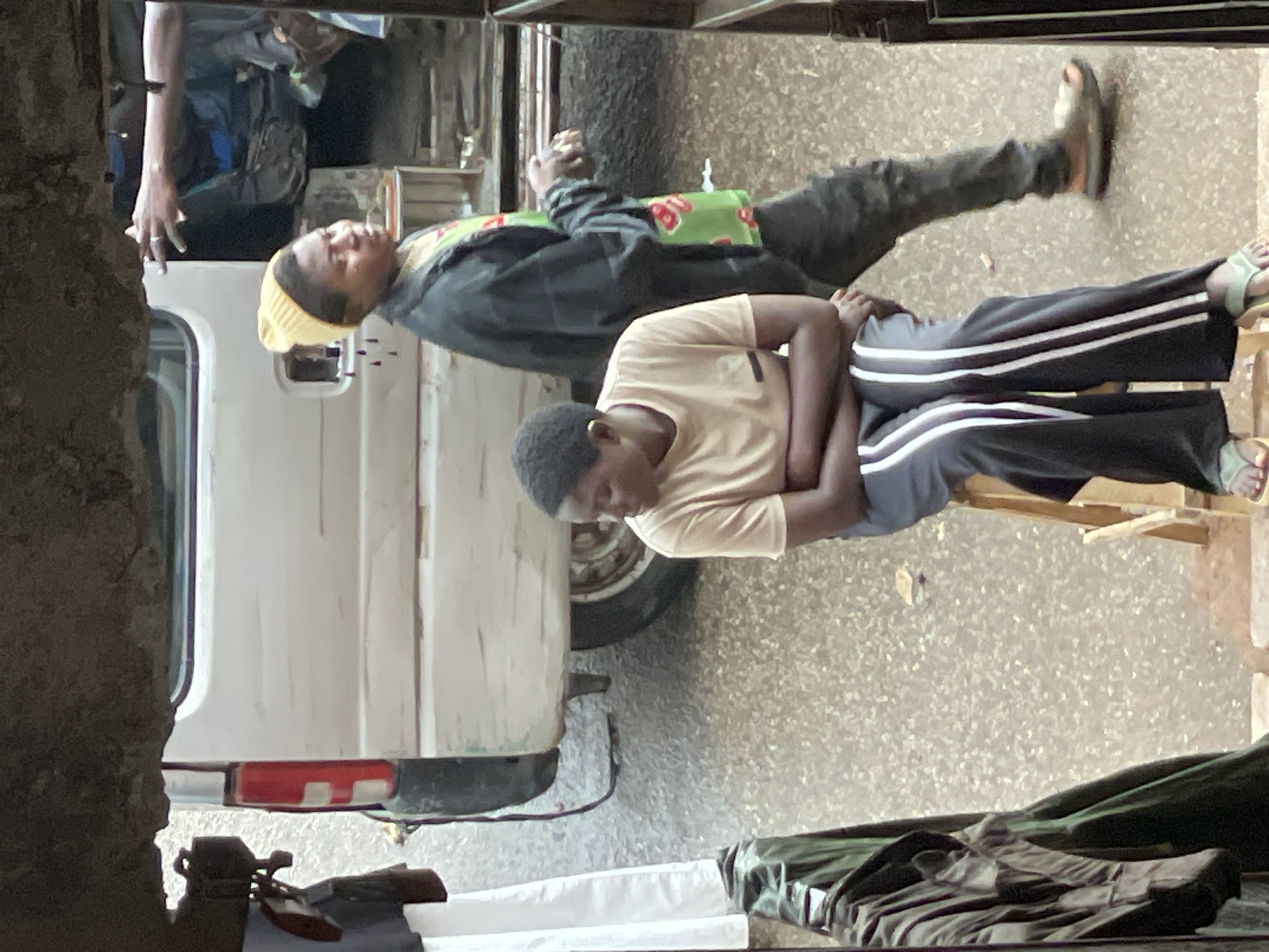 The image captures a scene in Bukuru, Nigeria. An adult male wearing a tan shirt and black pants with white stripes sits on a wooden chair, his arms crossed over his stomach, looking down. Behind him, another adult male wearing a yellow knit cap, a black jacket, and camouflage pants lies on a surface covered with a green and red patterned cloth. They are positioned near a white van. The ground appears to be concrete or paved. Items including clothing and possibly luggage lie on a surface beneath the van, suggesting a travel or temporary living situation. A concrete structure frames the left edge of the image, possibly part of a building, and the overall impression is that of an outdoor urban setting.