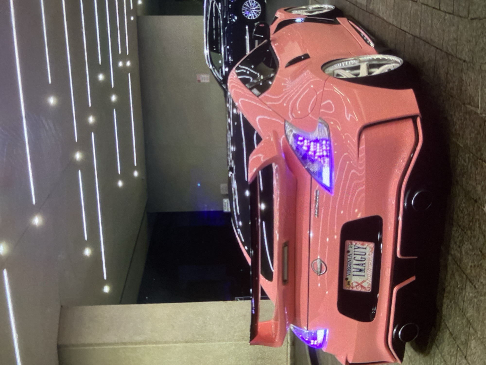 A close-up shot features the rear of a pink Nissan 370Z with a Virginia license plate that reads "IMAGUY". The car has aftermarket modifications, including a spoiler, taillights, and custom rear bumper with exhaust outlets. The scene appears to be in a parking garage, with artificial lighting visible in the background. Another dark-colored car is partially visible behind the Nissan. The image has no indication of location in Lhokseumawe, Indonesia.