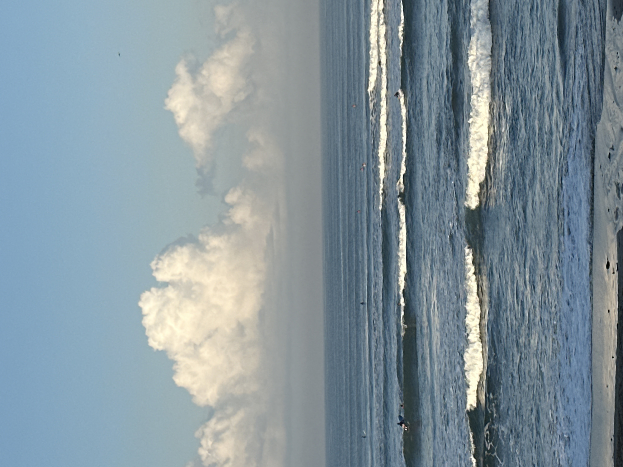 The image depicts a wide view of the ocean with several surfers in the water. The sky is bright blue with fluffy white clouds. The ocean has moderate waves, with several surfers riding them or paddling. In the distance, the horizon is blurred, suggesting a hazy or misty atmosphere. The foreground shows a strip of sand or beach. The location is identified as North Kuta, Indonesia. There are multiple individuals on surfboards scattered throughout the frame. Some are actively surfing on waves, while others are in a resting position on their boards. The overall scene conveys a typical day at the beach with surfing as the main activity.