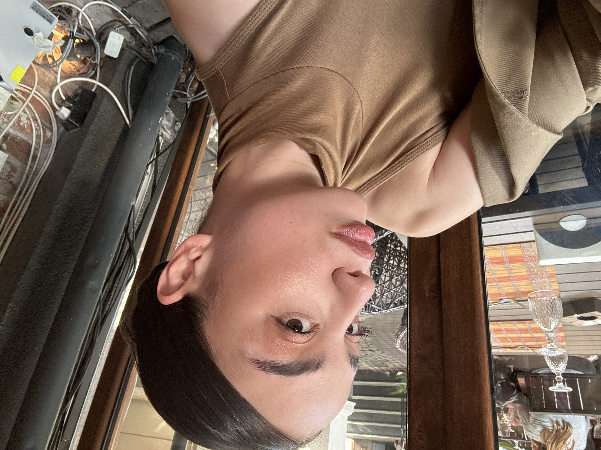 A woman wearing a brown top and jacket is visible. The setting appears to be an indoor space adjacent to an outdoor area. Visible in the background are electrical wires, brick, a black pipe, wooden window frames, and a reflective surface showing glasses on a table. The location is not possible to determine with the available data.